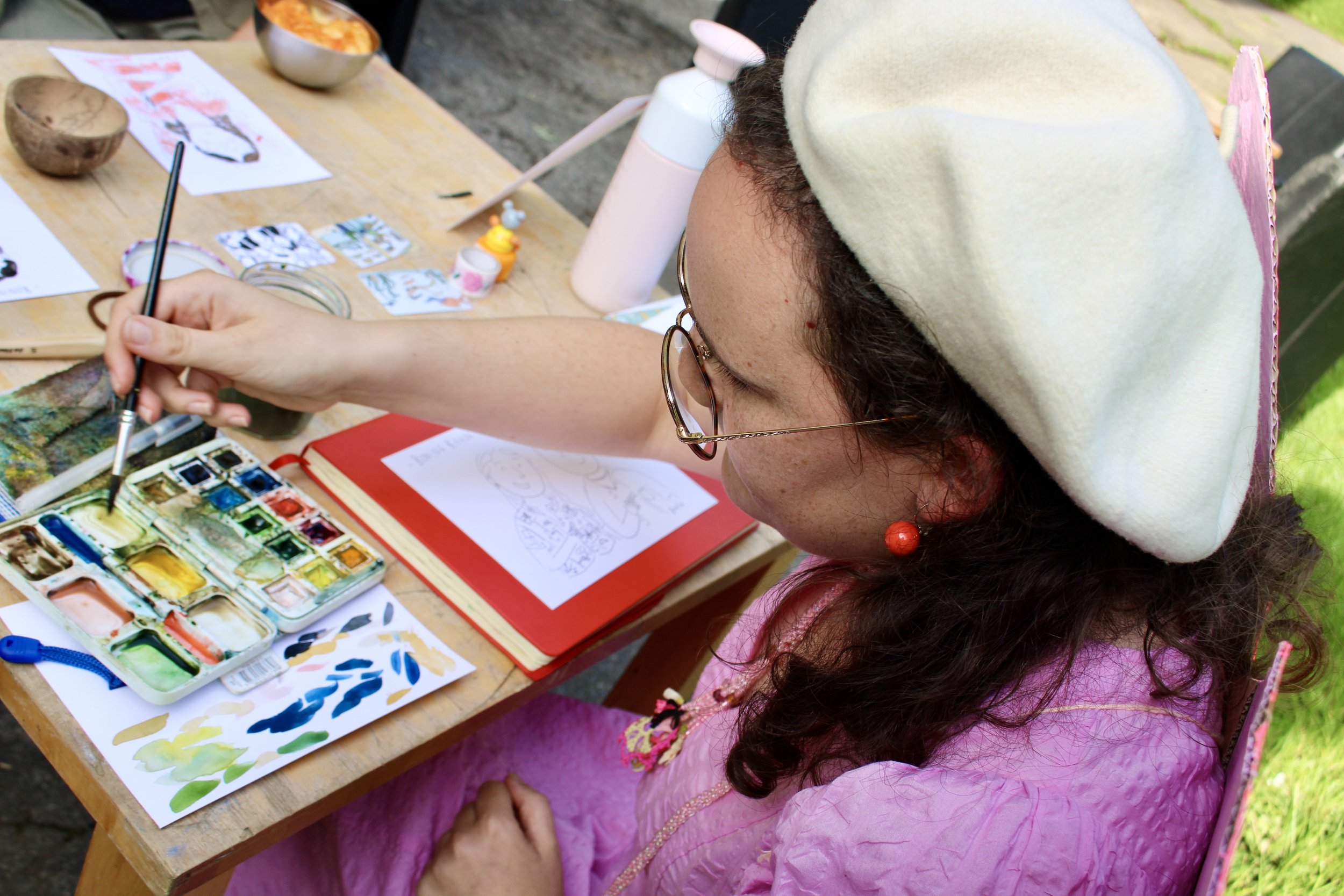 Vrouw in roze jurk en witte baret schildert met aquarellen aan tafel buiten