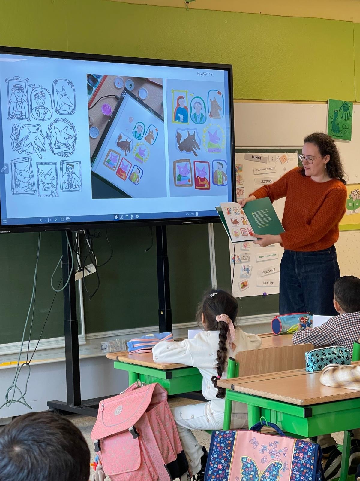 A classroom with a teacher presenting something on a big screen. There are children sitting on their chairs watching the presentation. The screen shows coloring pages and drawings. The children have their school supplies and bags on their desks.