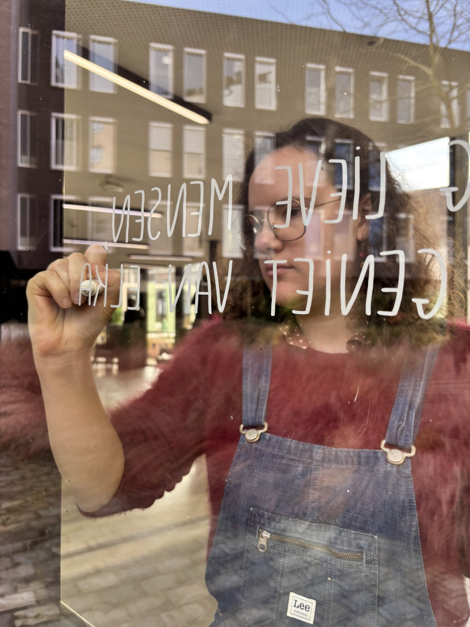 Person writes with marker on a glass door, text reflected in the glass. Building and street visible in the reflection.