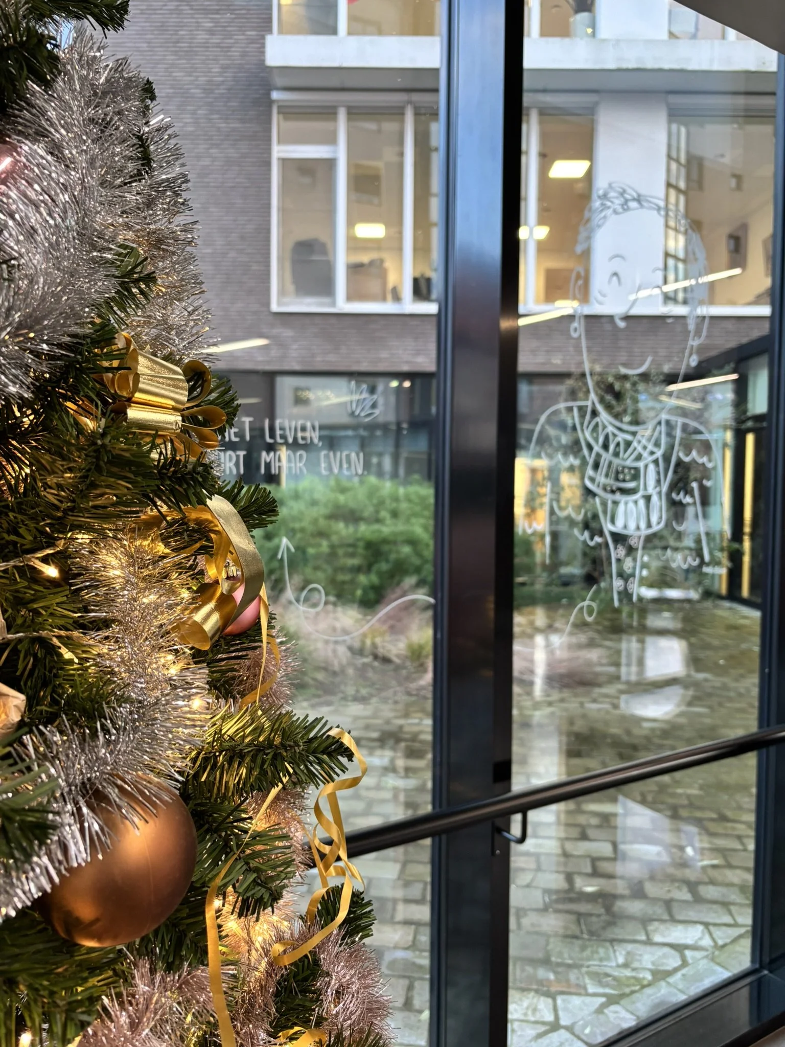 Christmas tree decoration with gold and pink balls and ribbons in front of a glass door with drawing on the window, view of building across the street.