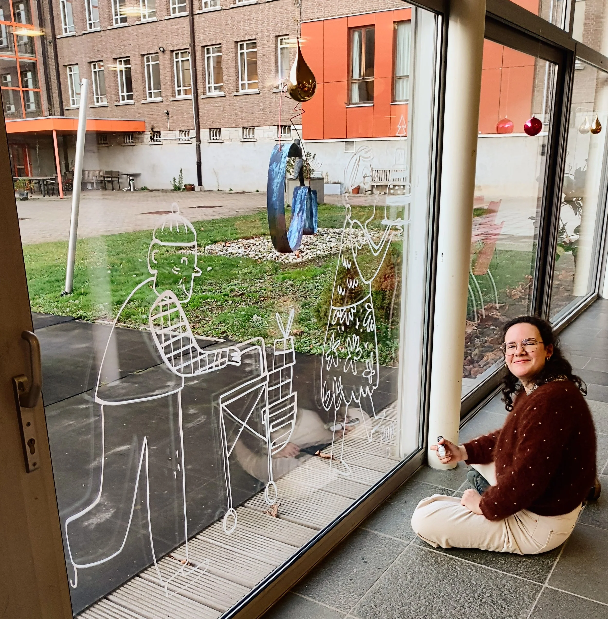 Aurélie est assise par terre à côté d'une paroi en verre avec des illustrations dessinées, dont une personne avec un déambulateur et quelqu'un jouant de la trompette ; vue extérieure de la pelouse et de la sculpture.