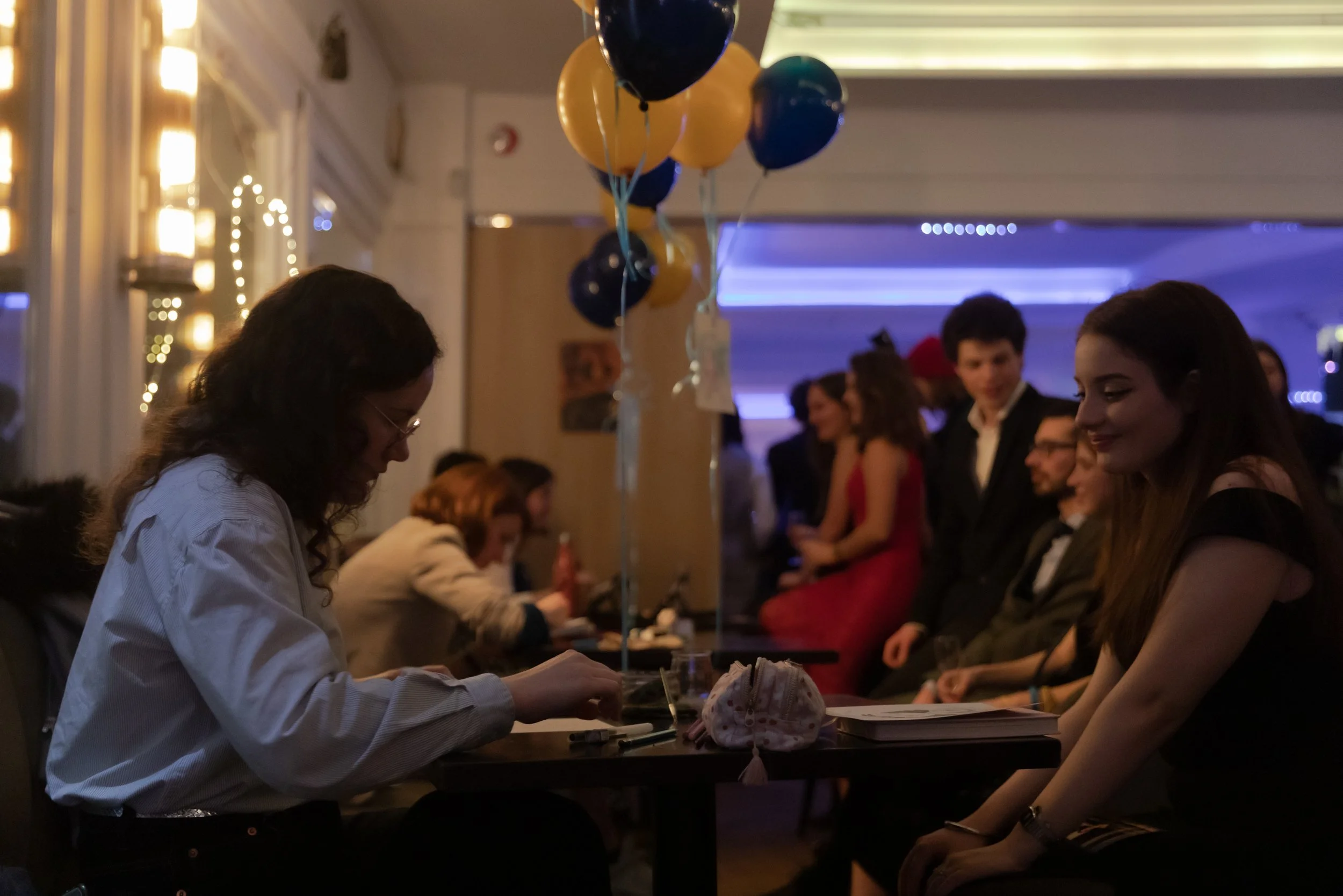 Personnes dans un restaurant ou un café, assises à des tables, certaines en tenue de soirée ; ballons en arrière-plan.