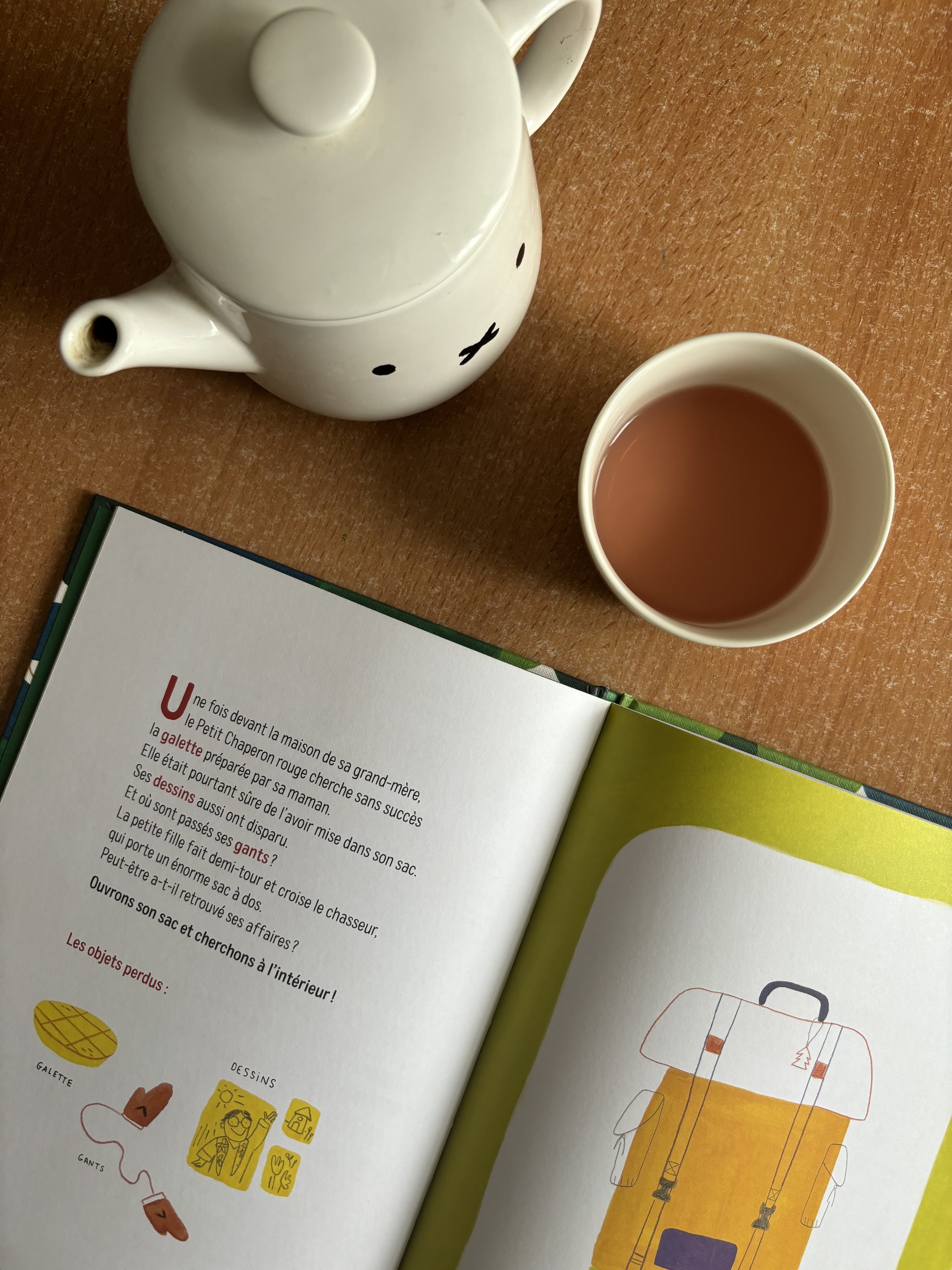 White teapot with face, cup with tea, and open book with illustrations on wooden table.