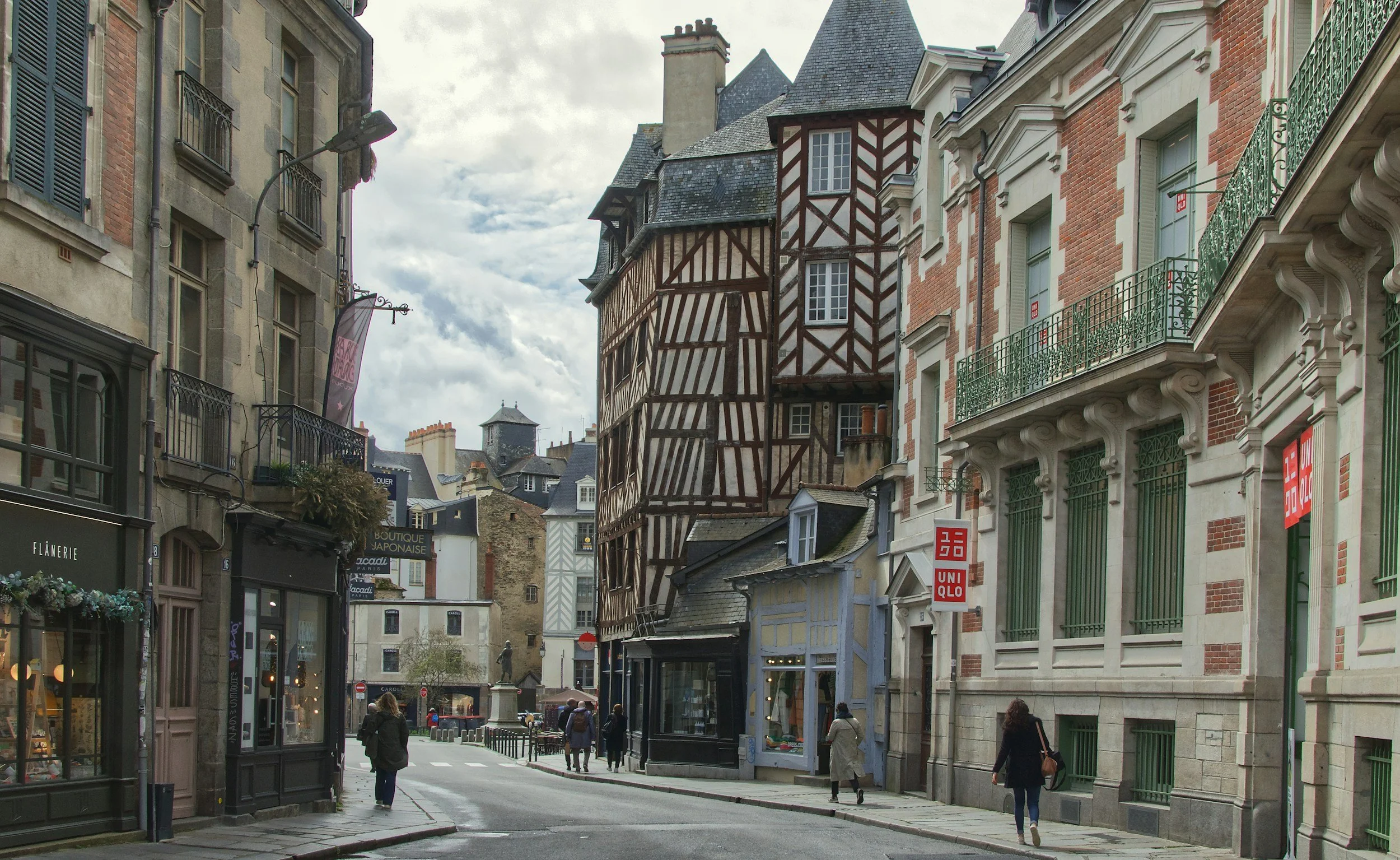 A narrow cobblestone street in a historic European town with stone buildings, flowering bushes, and a few pedestrians enjoying a sunny day.