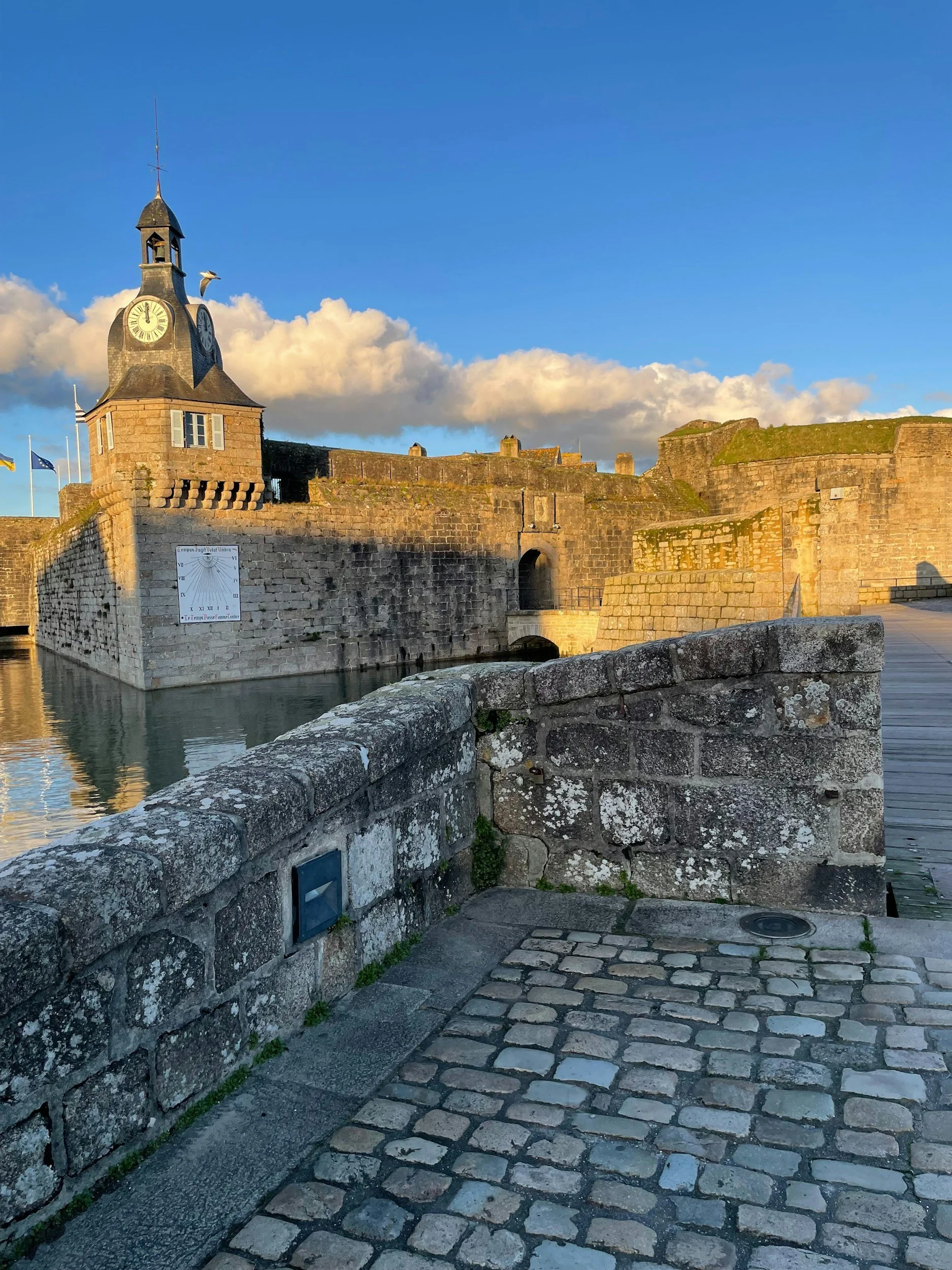 Historical courtyard with a stone fountain, cobblestone ground, and multi-story buildings with shuttered windows and wrought iron balconies, under a blue sky.