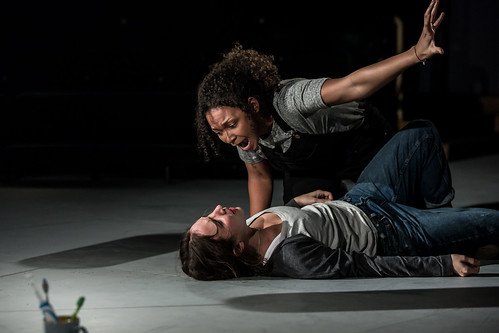 A young white woman with brown hair, jeans and white top lies on a mostly bare stage as another young, black woman in navy dungarees leans over her shouting.