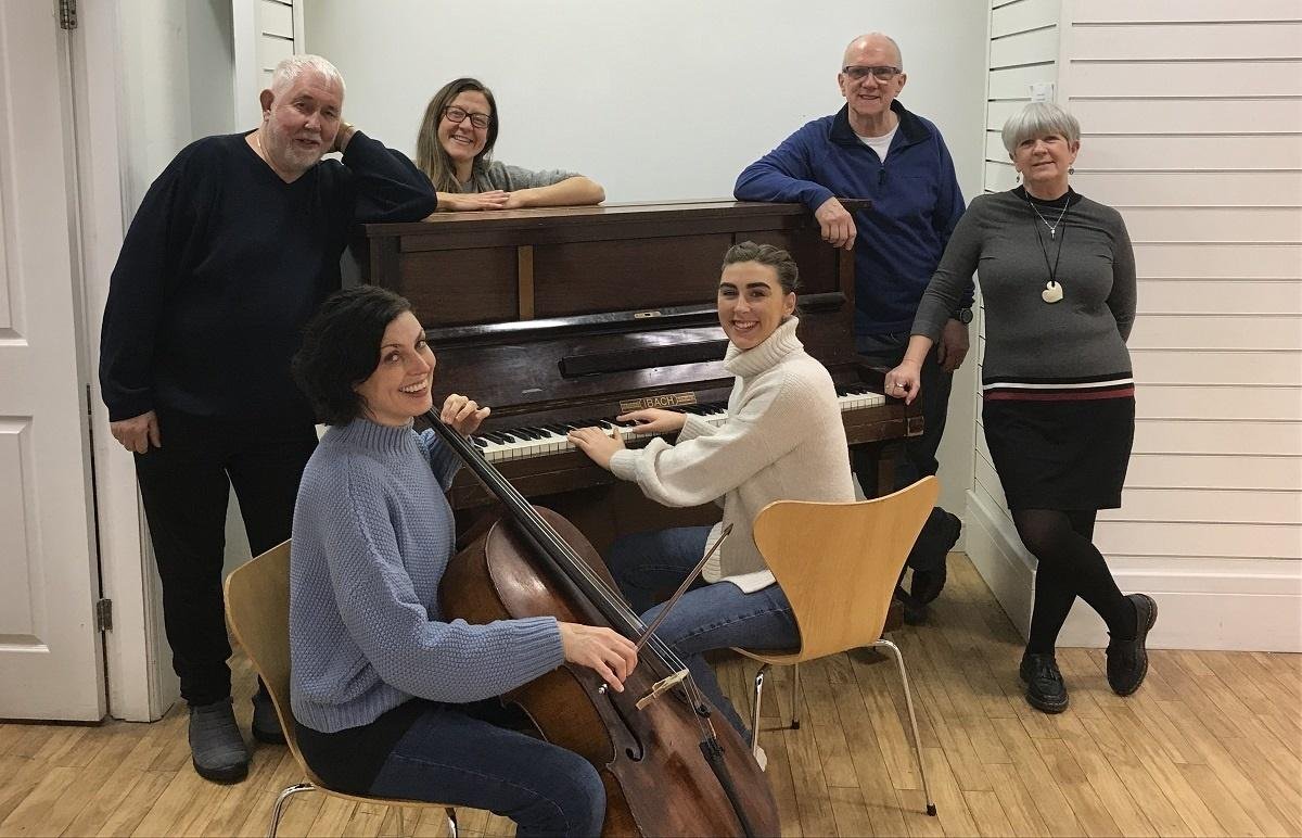 Four white women and two white men stand around a piano smiling. The woman at the front is also holding a cello.
