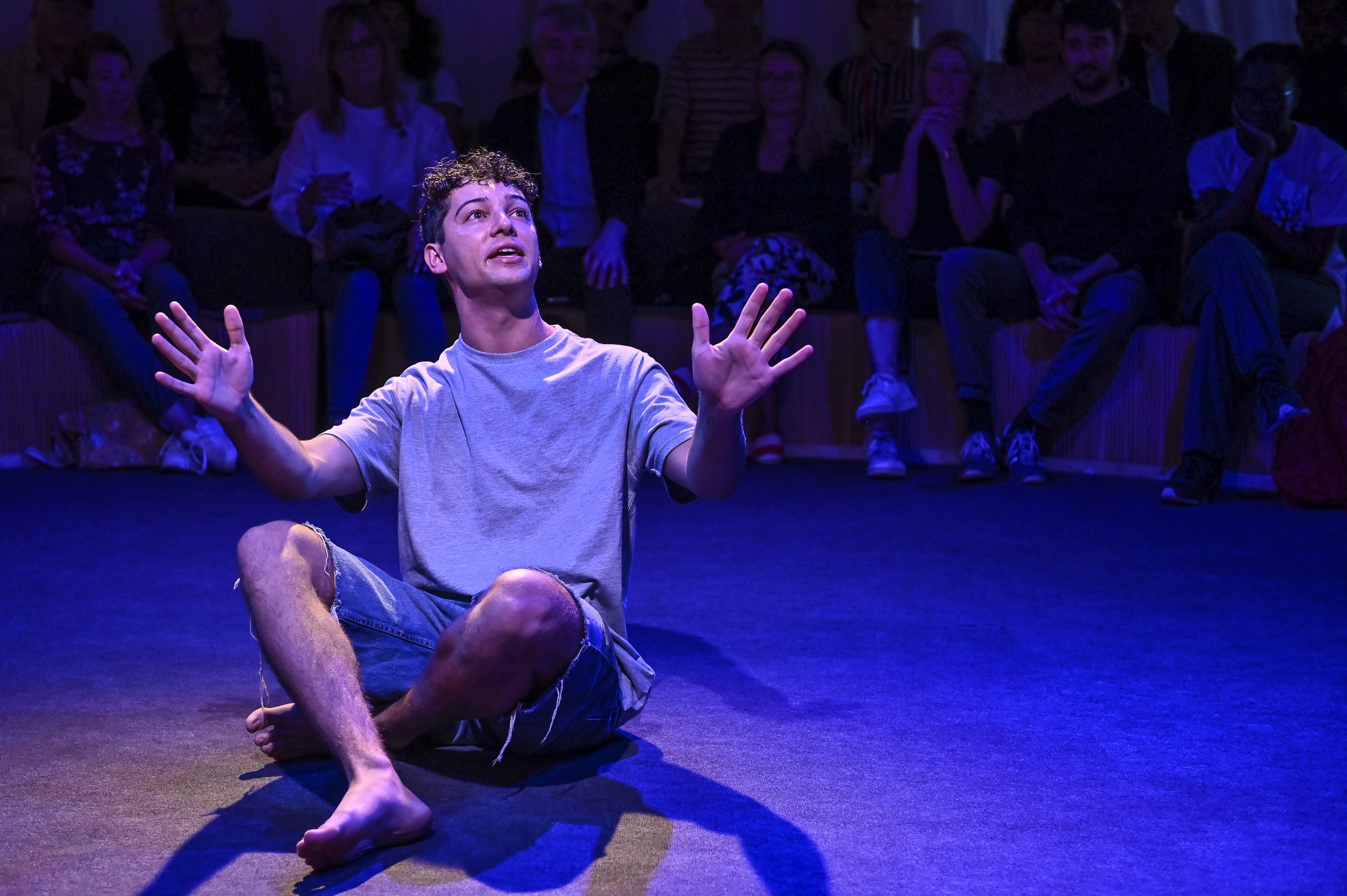 A young man sits on stage in purple light, his hands reach out to illustrate his story.