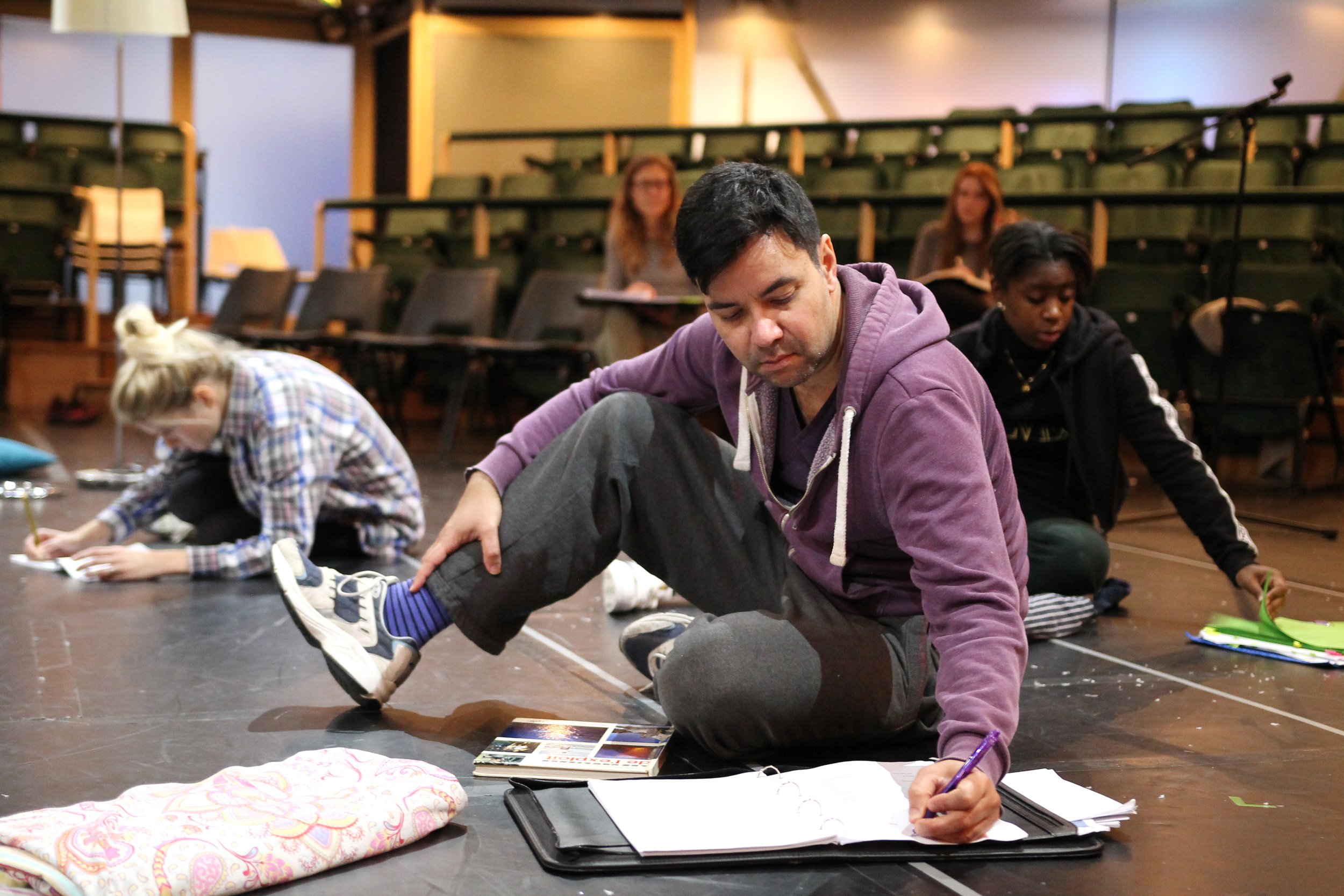An East Asian man, white woman and black woman sit on stage looking at scripts in folders. In the background are empty theatre seats with two seated white women barely visible.