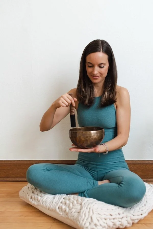 A white woman with brown hair wearing blue yoga clothes is sitting on a white pillow with her legs crossed, playing a brass bowl held in her palm at chest height.