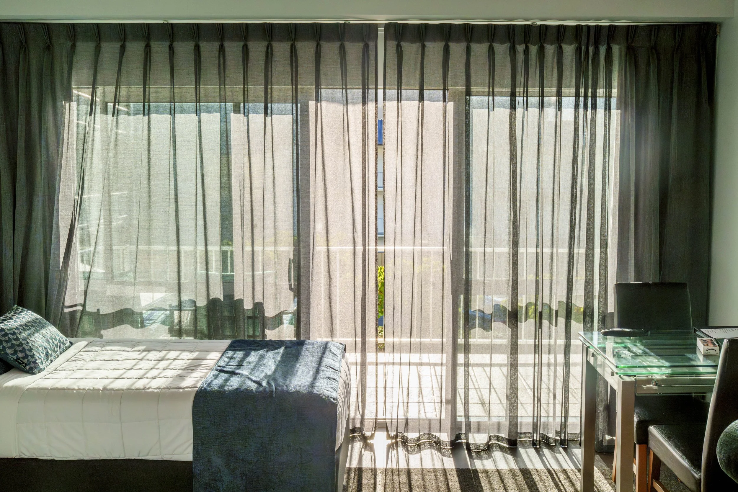 View of a bedroom with large sliding glass doors covered by sheer curtains, a bed on the left with pillows and a blanket, and a glass-topped table on the right with some items on it.