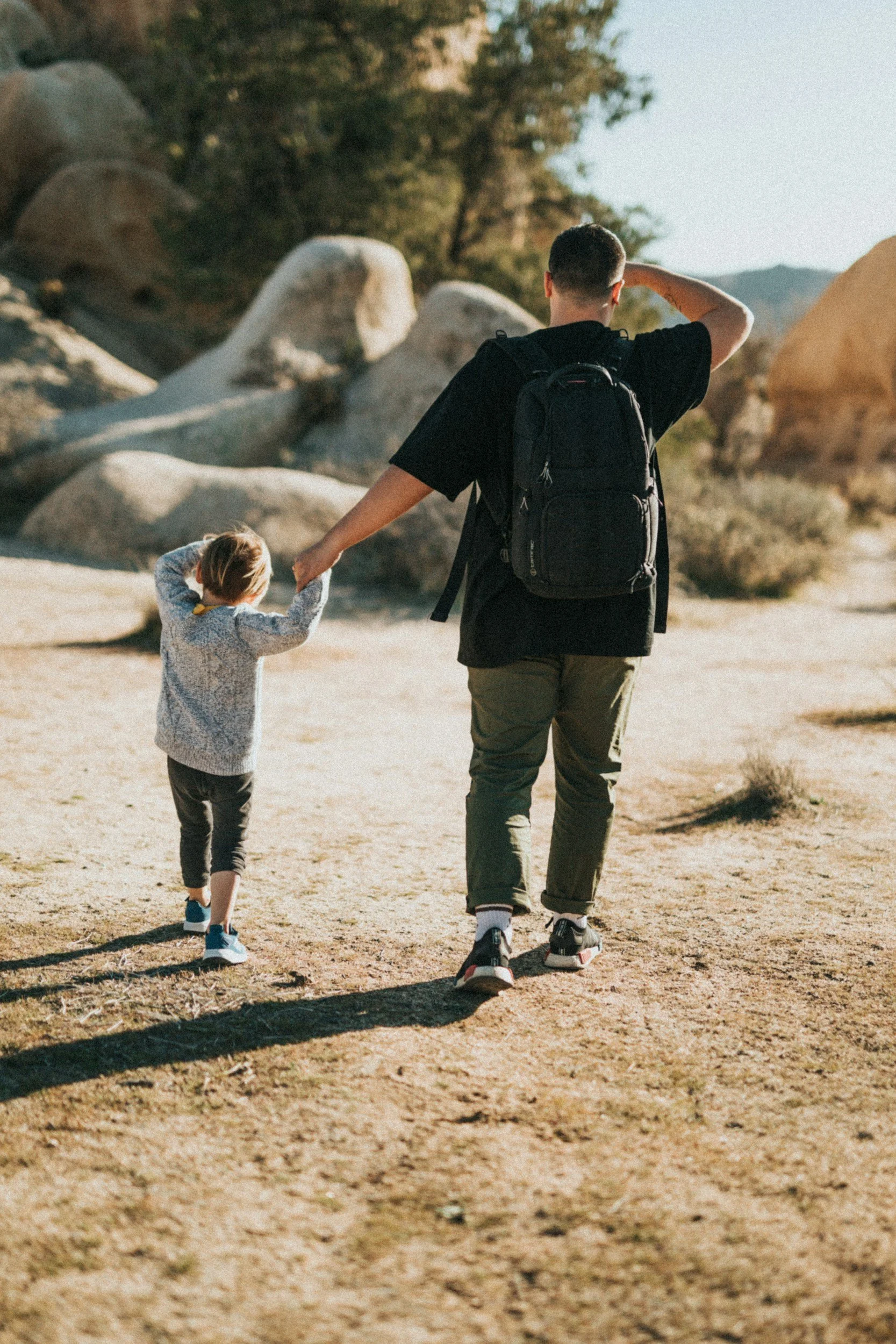 A man and a young girl walking hand in hand on a dirt trail in a rocky desert landscape with trees, under a sunny sky.