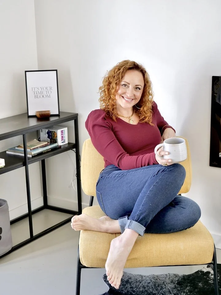 A woman with curly red hair sitting cross-legged on a yellow armchair, holding a gray mug and smiling at the camera. She is in a modern living room with a black bookshelf and a framed motivational quote, 'It's your time to bloom,' on a side table.