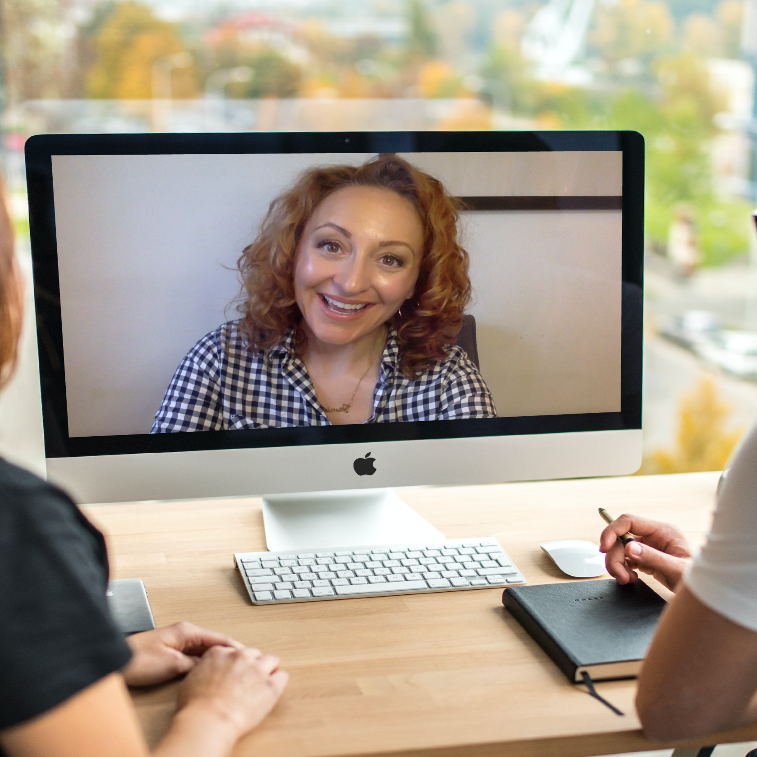 Woman on computer screen smiling during a video call, sitting at a desk with two people in front of her, one writing in a notebook, in a bright room with a window showing trees outside.