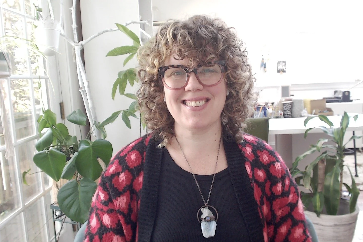 A woman with curly blonde hair, glasses, and a red and black patterned sweater smiling at the camera, sitting indoors with plants and a bright window behind her.