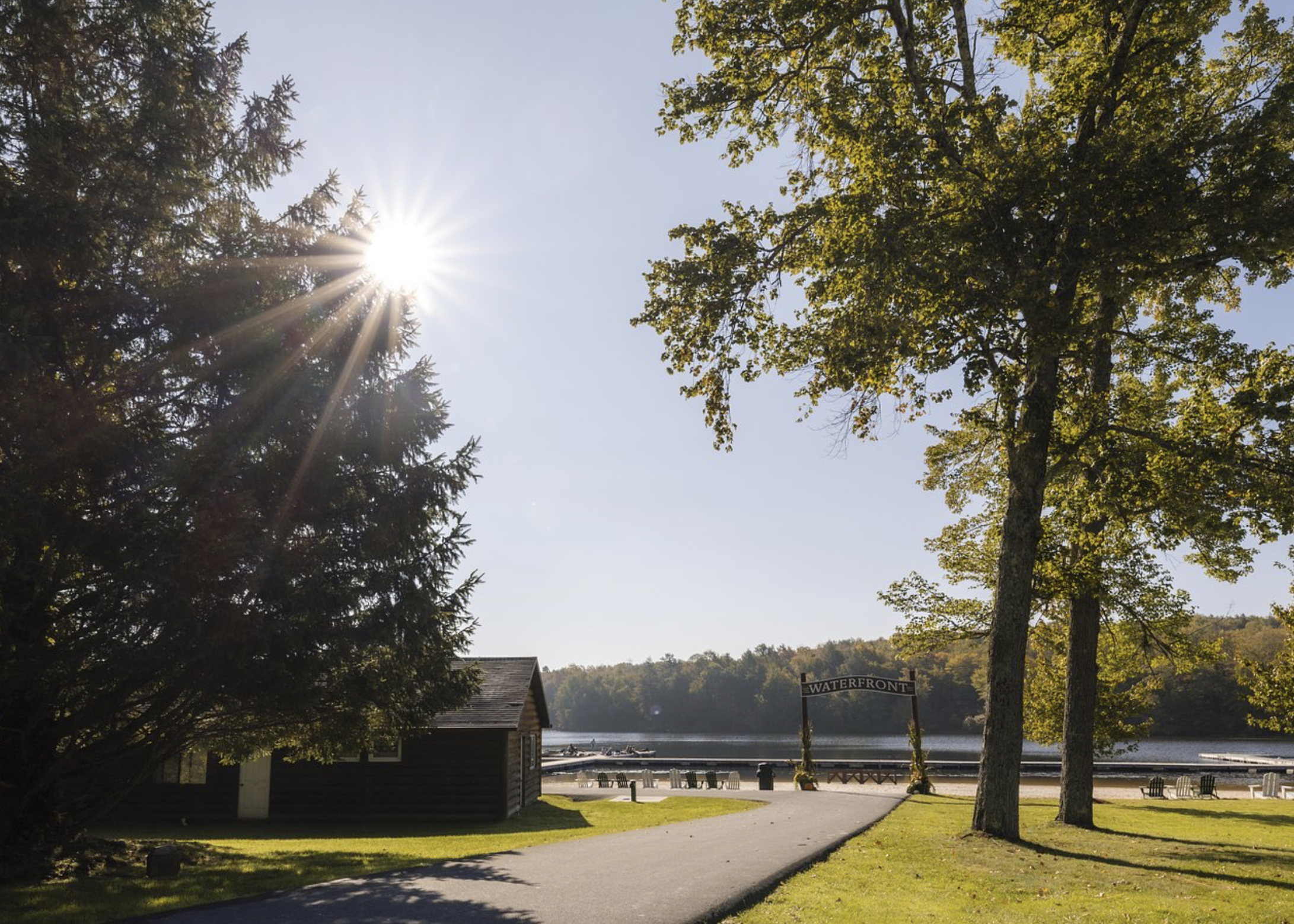Sun shining through trees over water and waterfront area with benches, chairs, and a small building, with a sign that reads 'Waterfront'.
