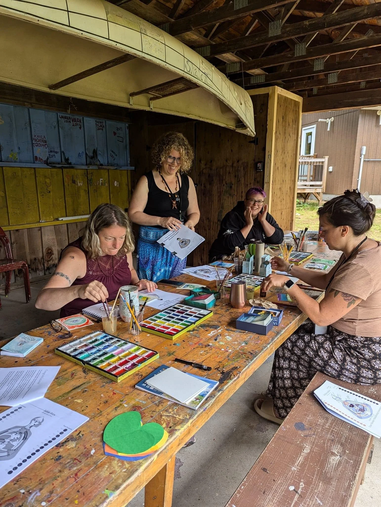 Group of women at a craft workshop sitting at a wooden table with watercolor paints, brushes, and craft supplies. One woman stands smiling while holding papers, others are engaged in arts and crafts in an outdoor covered area with wooden walls and roof.