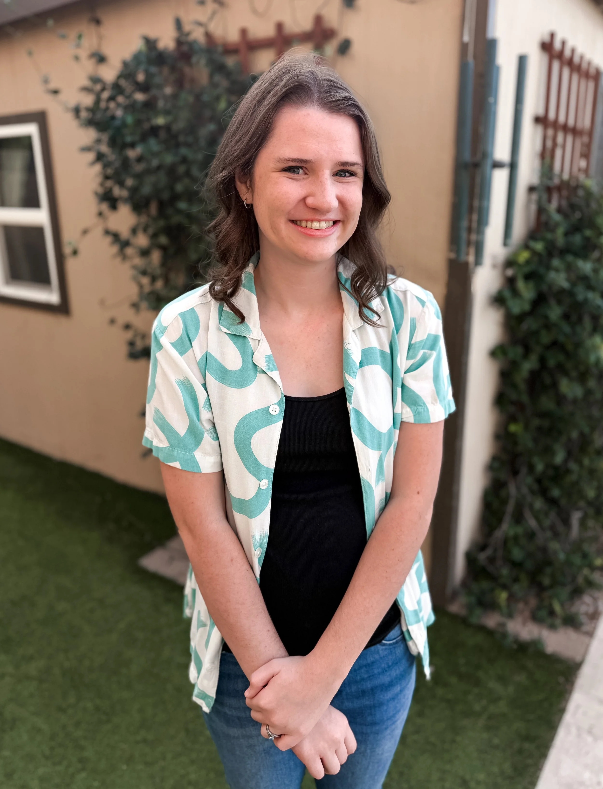 Berlynn Burns from The  Burns Creative standing outdoors, smiling, wearing a black top and a white short-sleeved shirt with teal abstract patterns, standing on a patch of green grass with a beige house and greenery in the background.