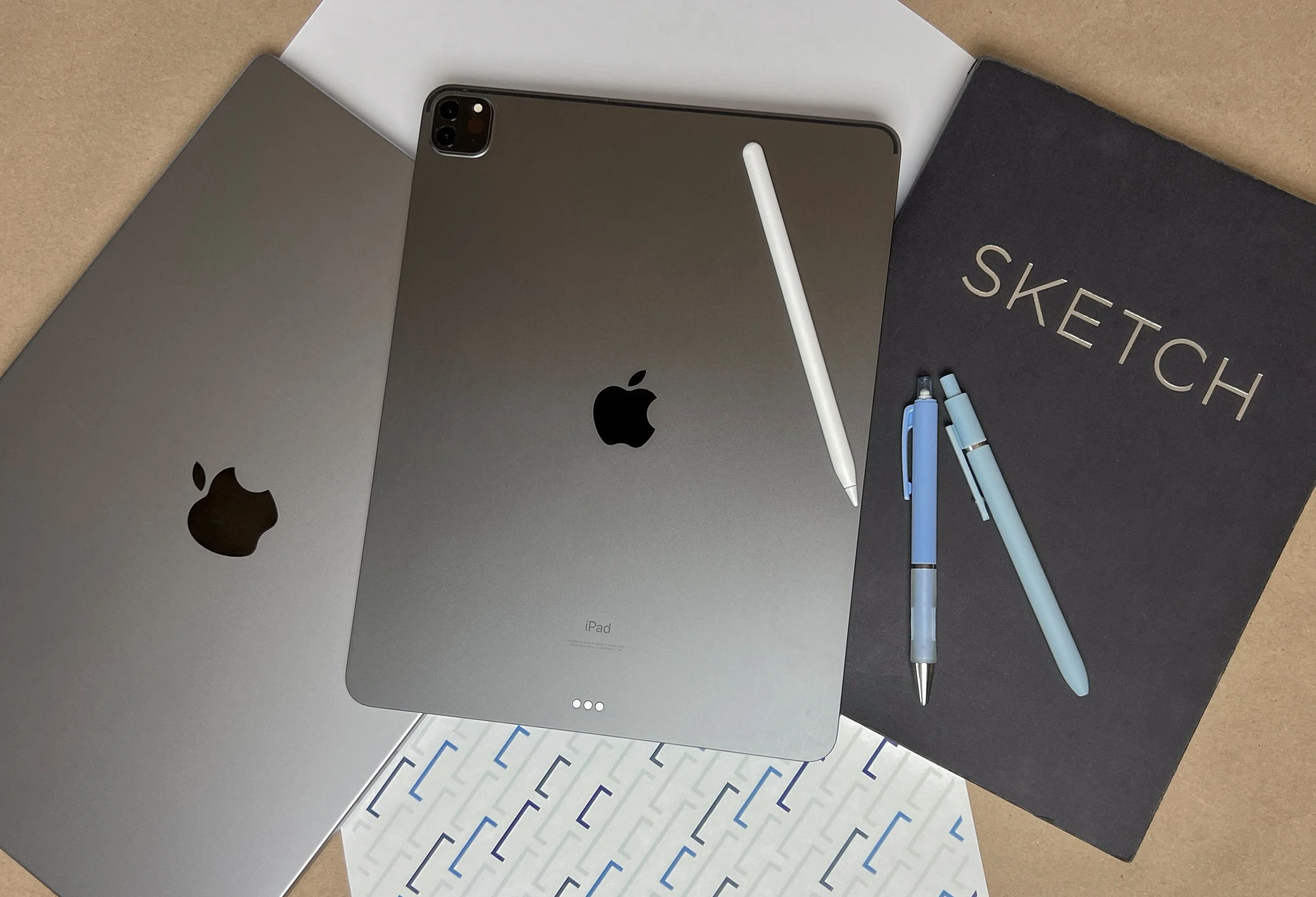 Top view of Berlynn Burns' workspace with two MacBook laptops, an iPad with a stylus, two blue pens, a black sketchbook with white text, and a piece of patterned paper. The tools used by The Burns Creative