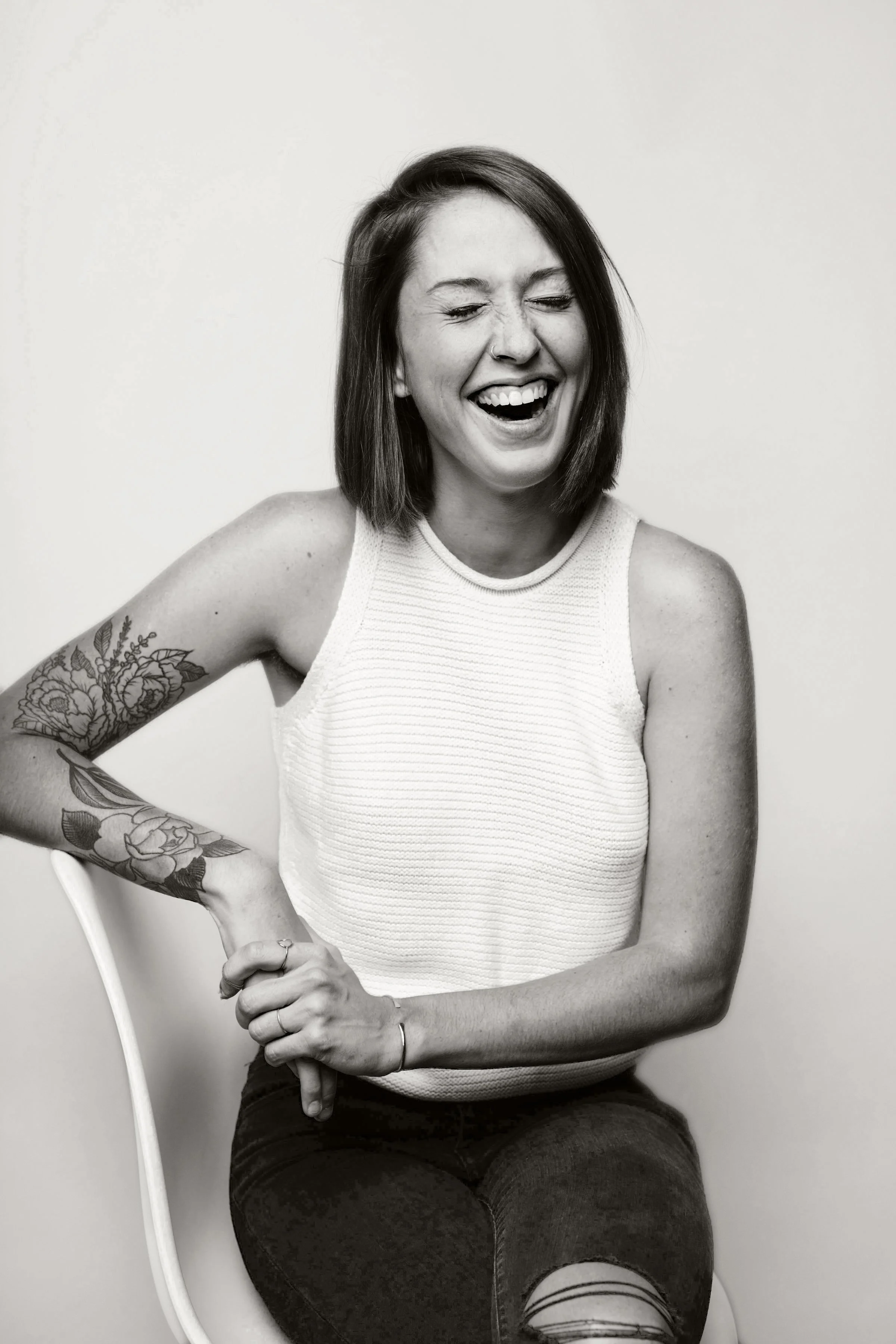 A young woman with short dark hair and tattoos on her left arm, smiling and laughing with her eyes closed, seated on a chair against a plain background.