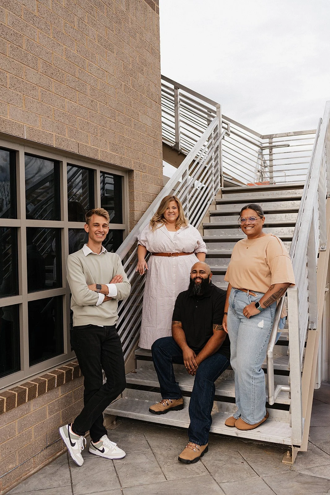 A diverse group of five people, three women and two men, smiling and posing on an outdoor metal staircase with a brick building in the background.