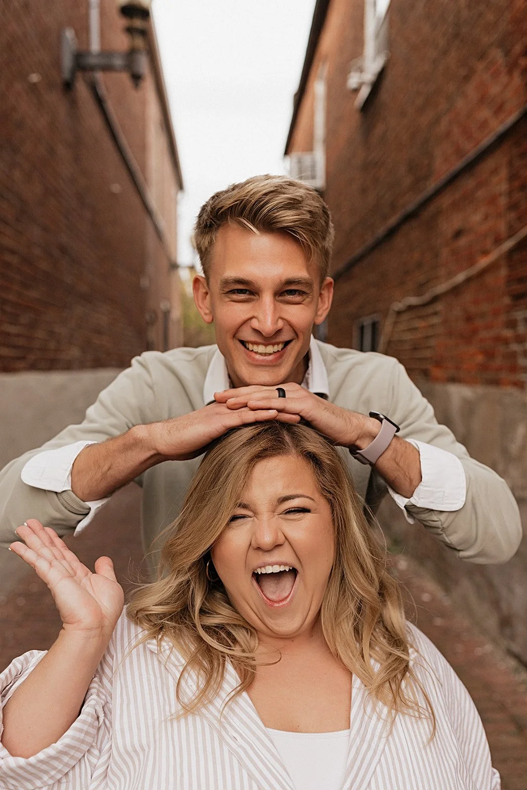 A young man and older woman playfully smiling and laughing in an alleyway between brick buildings, with the man placing his hands on the woman's head.