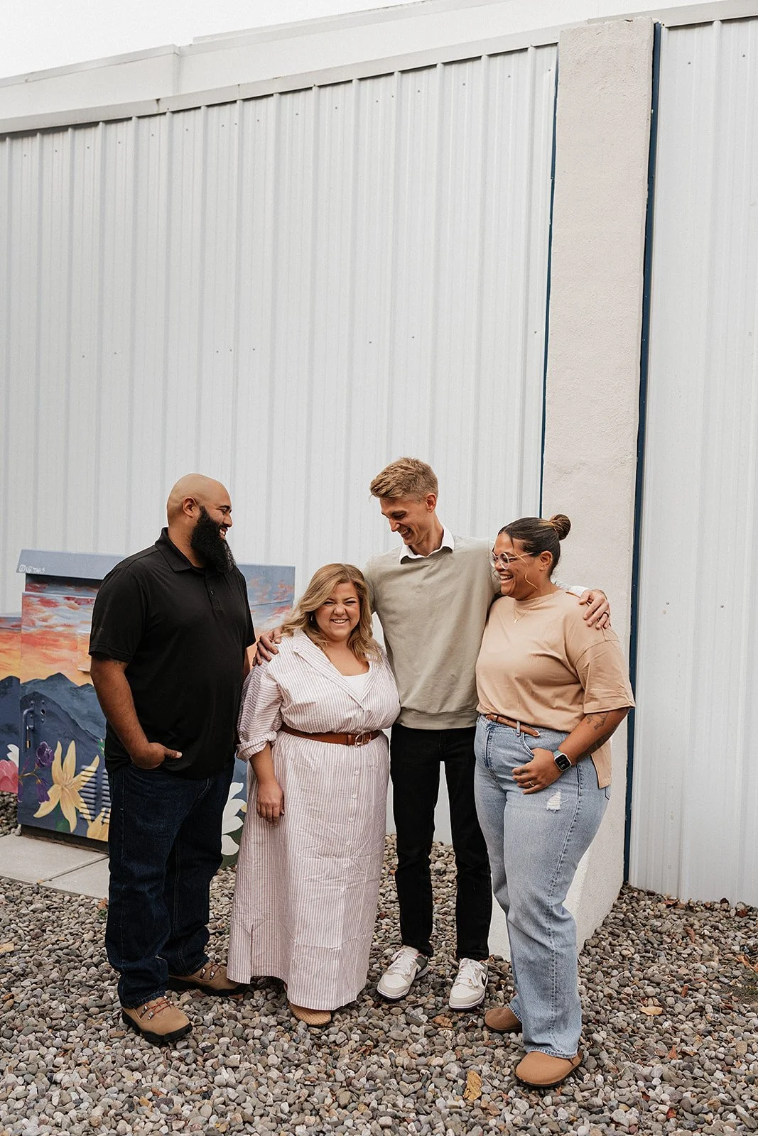 Group of five diverse people smiling and standing together outdoors in front of a metal building and painted mural, on a gravel surface.