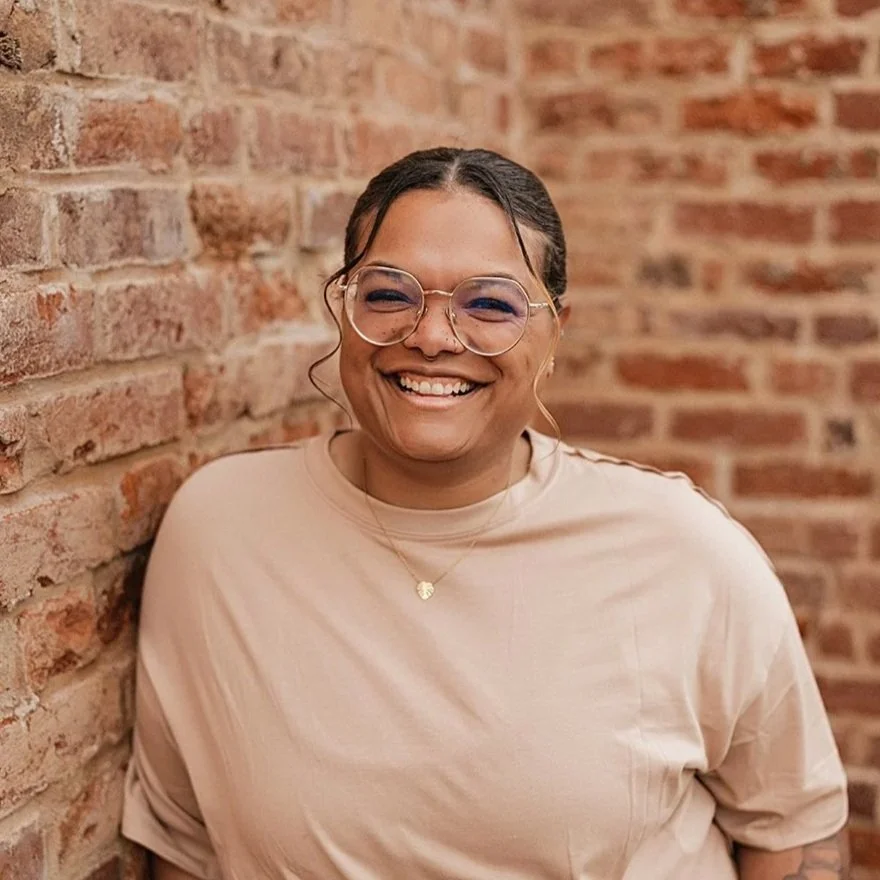 A woman with glasses, smiling, wearing a beige top, standing against a brick wall.