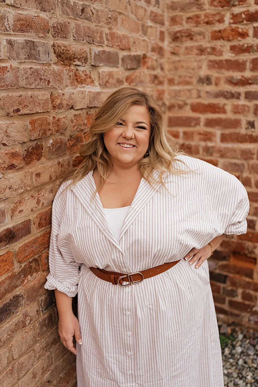 A woman with wavy blonde hair smiling and winking, wearing a white and beige striped button-down dress with rolled-up sleeves and a brown belt, standing against a brick wall.