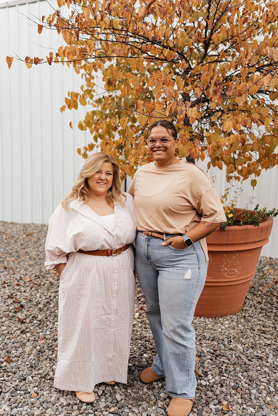 Two women standing outdoors in front of an orange autumn tree, smiling, casually dressed, with a large terracotta planter behind them.