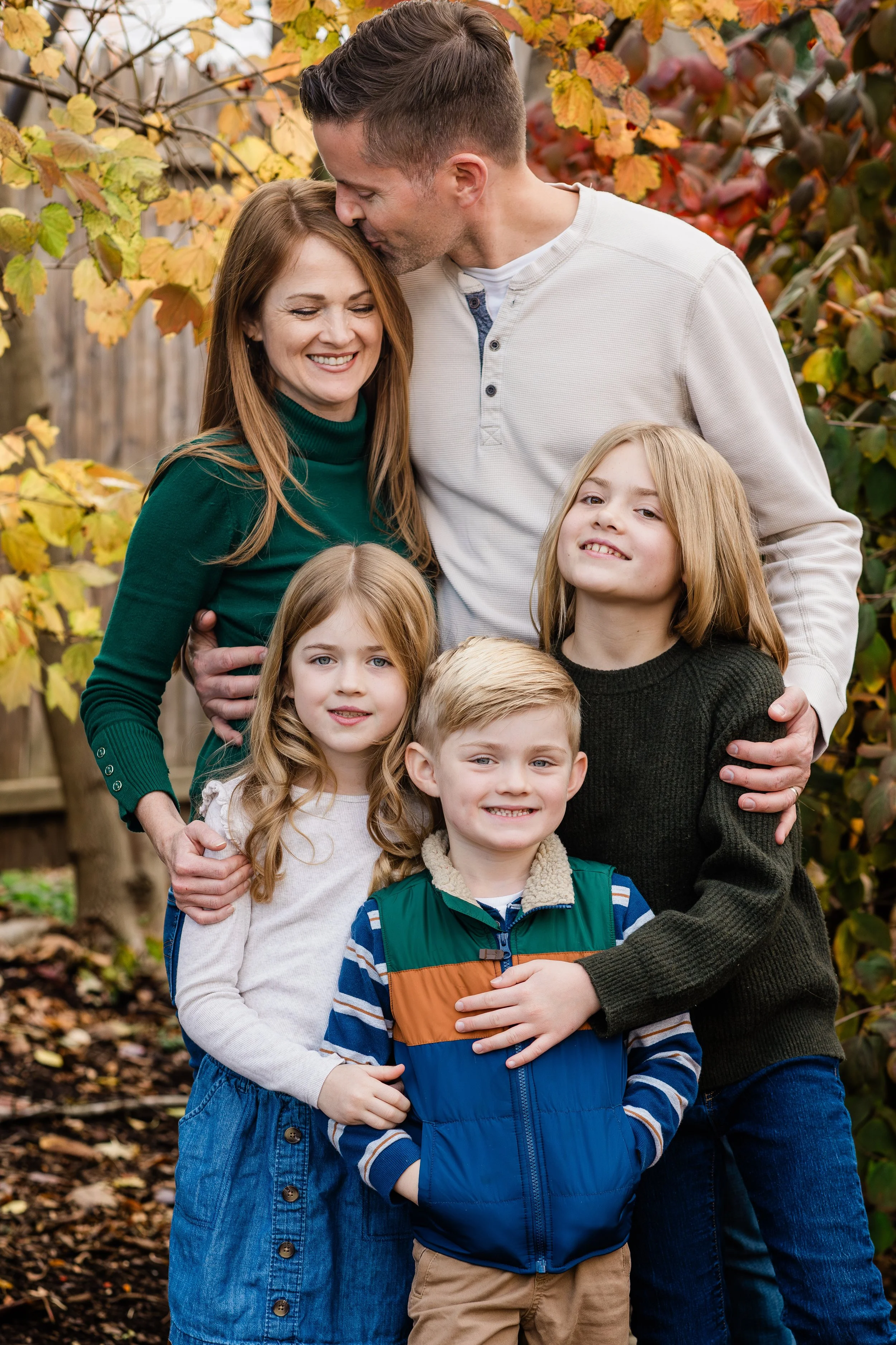 A family of six posing outdoors in autumn, surrounded by fall foliage. The mother and father are standing behind their three children, with the mother wearing a green turtleneck and the father in a light-colored long sleeve shirt. The children are a 