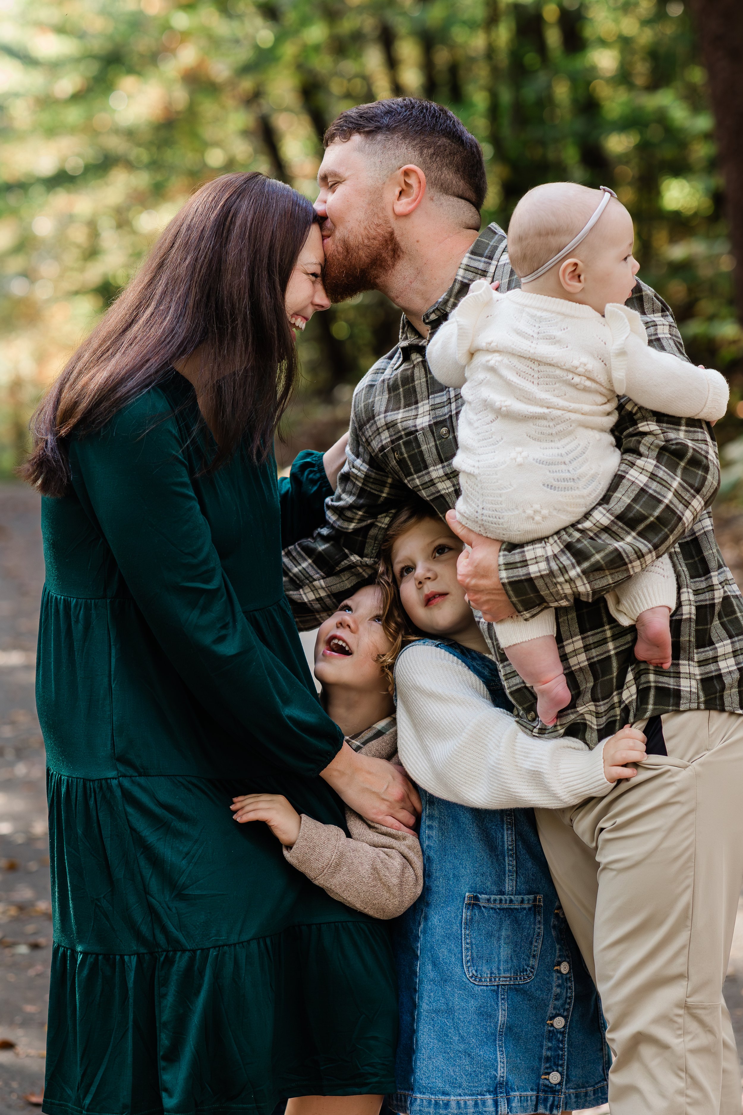 A family of five in a forest, with a man kissing a woman on the forehead, children hugging and looking up, and a baby in the man's arms.