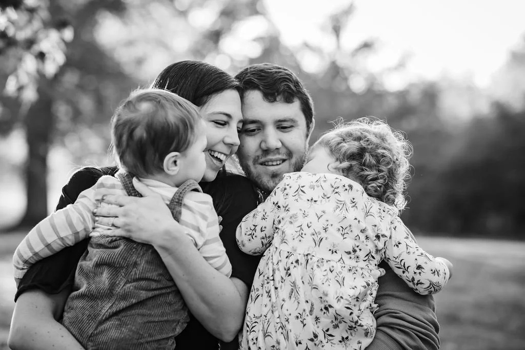 A black and white photo of a happy family embracing outdoors, with a woman, man, and two young children.