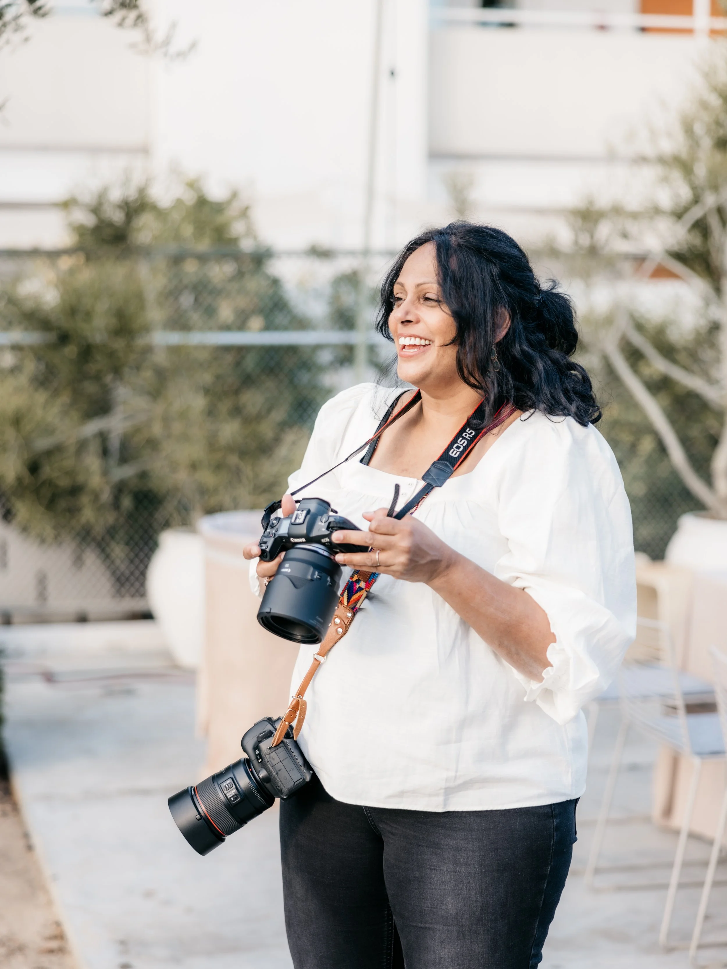 Smiling woman with black hair holding two professional cameras outdoors.