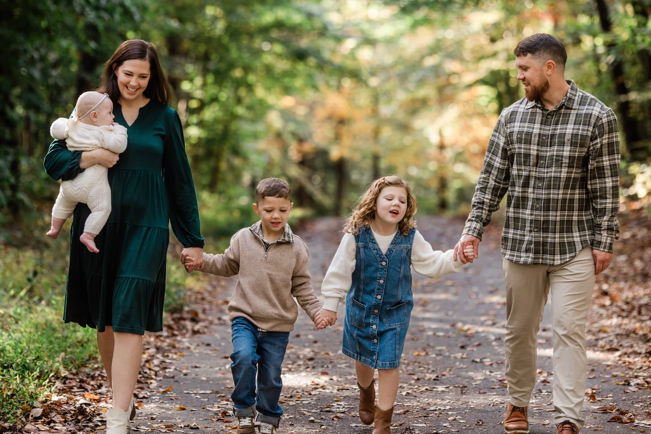 A family of six walking on a forest trail during fall, holding hands and smiling, with colorful autumn leaves on the ground.