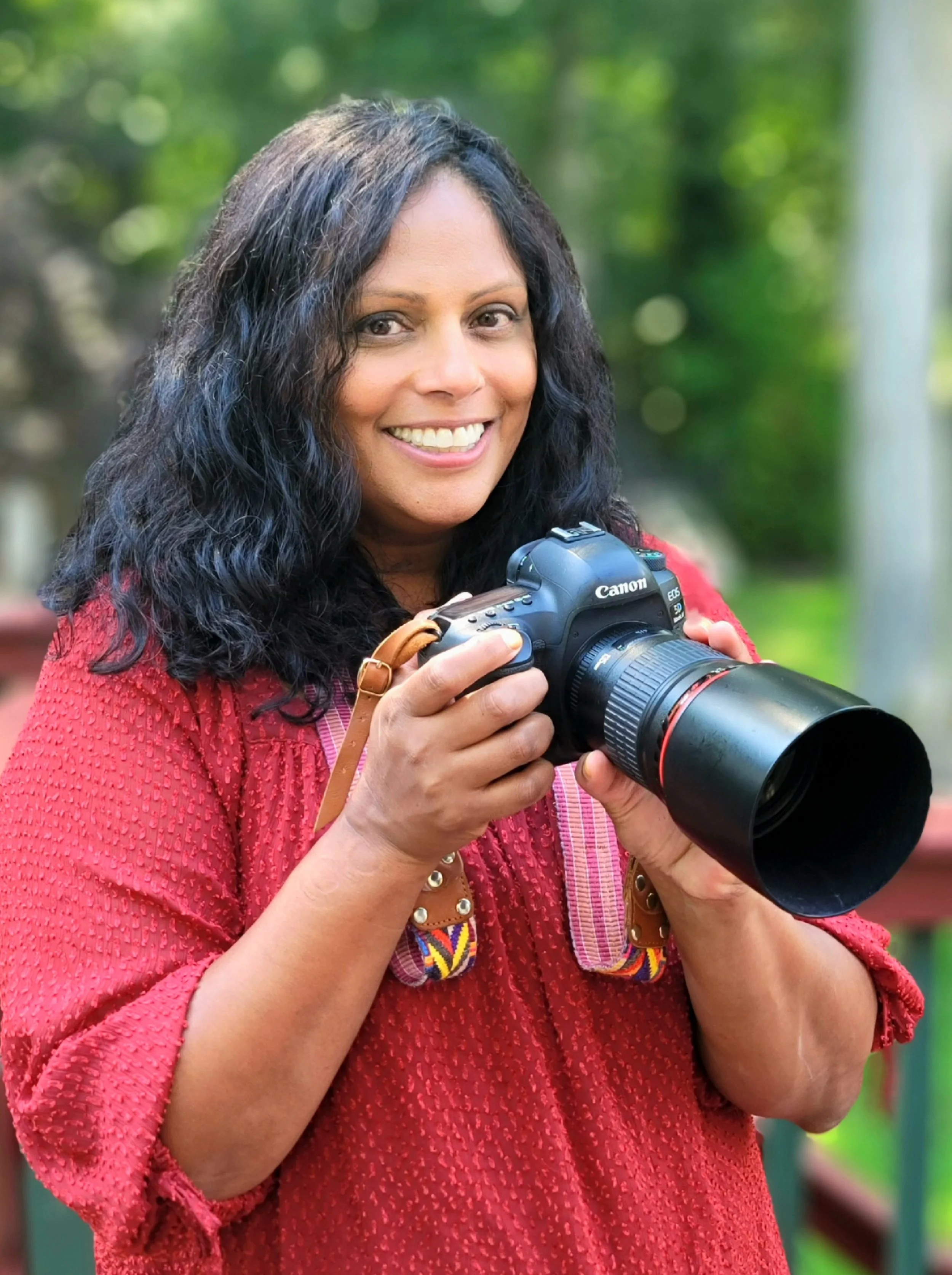 A woman with long, curly black hair holding a professional camera with a large zoom lens, smiling outdoors with a blurred background of green trees.