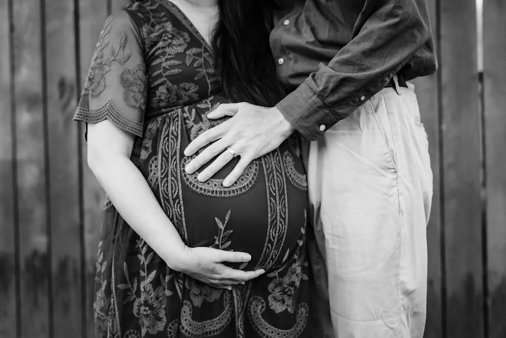 A pregnant woman in a floral-patterned dress and a man with a dark shirt and light-colored pants holding her belly and chest, standing in front of a wooden fence.