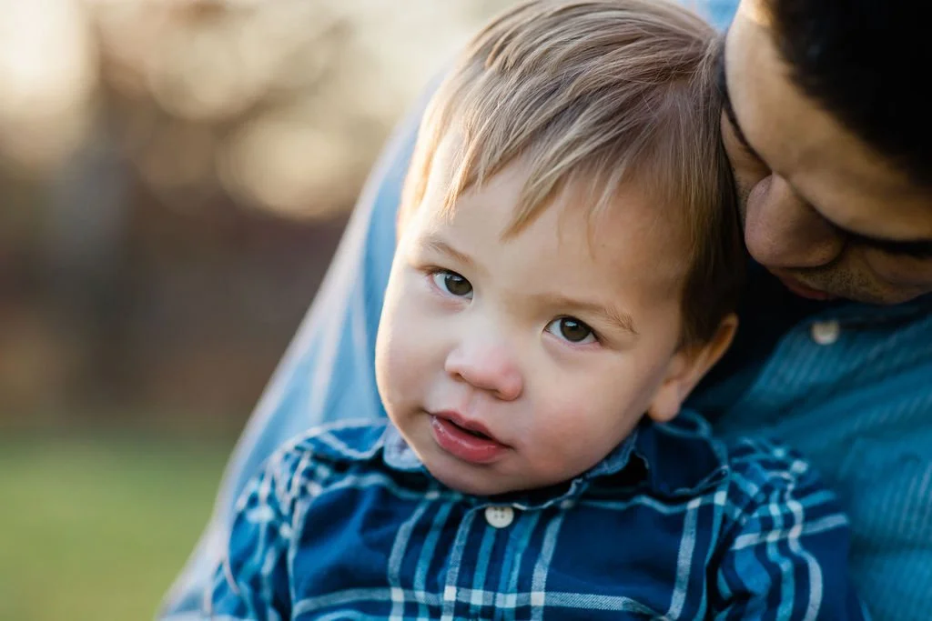 A young boy with light brown hair and blue eyes being held closely by an adult man, outdoors during daytime.