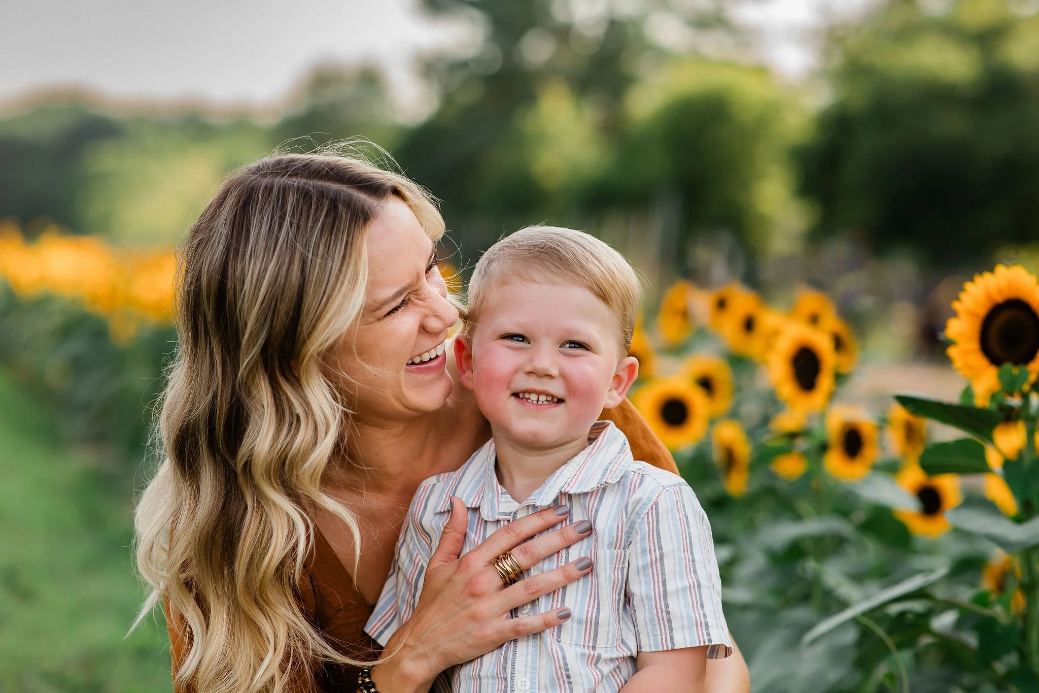 A woman and a young boy smiling and hugging in a sunflower field during daytime.