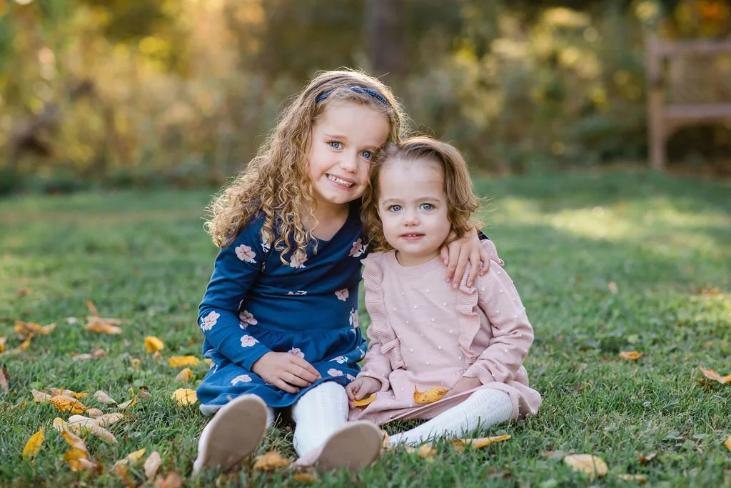 Two young girls sitting on the grass in a park during fall, with yellow leaves scattered around. The older girl has curly blonde hair, wearing a blue dress with pink and white flowers. The younger girl has light brown hair, wearing a light pink dress