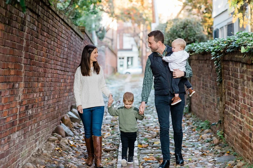 A happy family of four walking together on a rocky alleyway between brick walls during autumn, holding hands and smiling at each other.