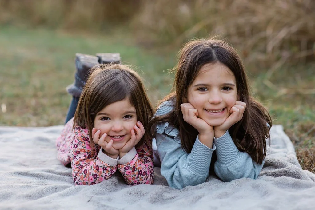 Two young girls lying on their stomachs on a blanket outdoors, smiling at the camera.