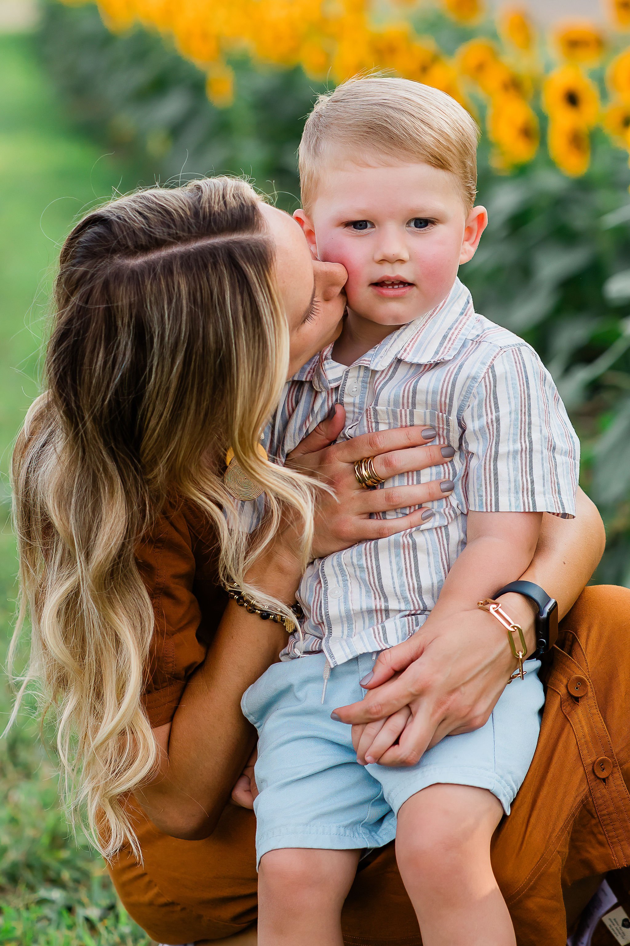 A woman kisses a young boy on the cheek in a sunflower field.