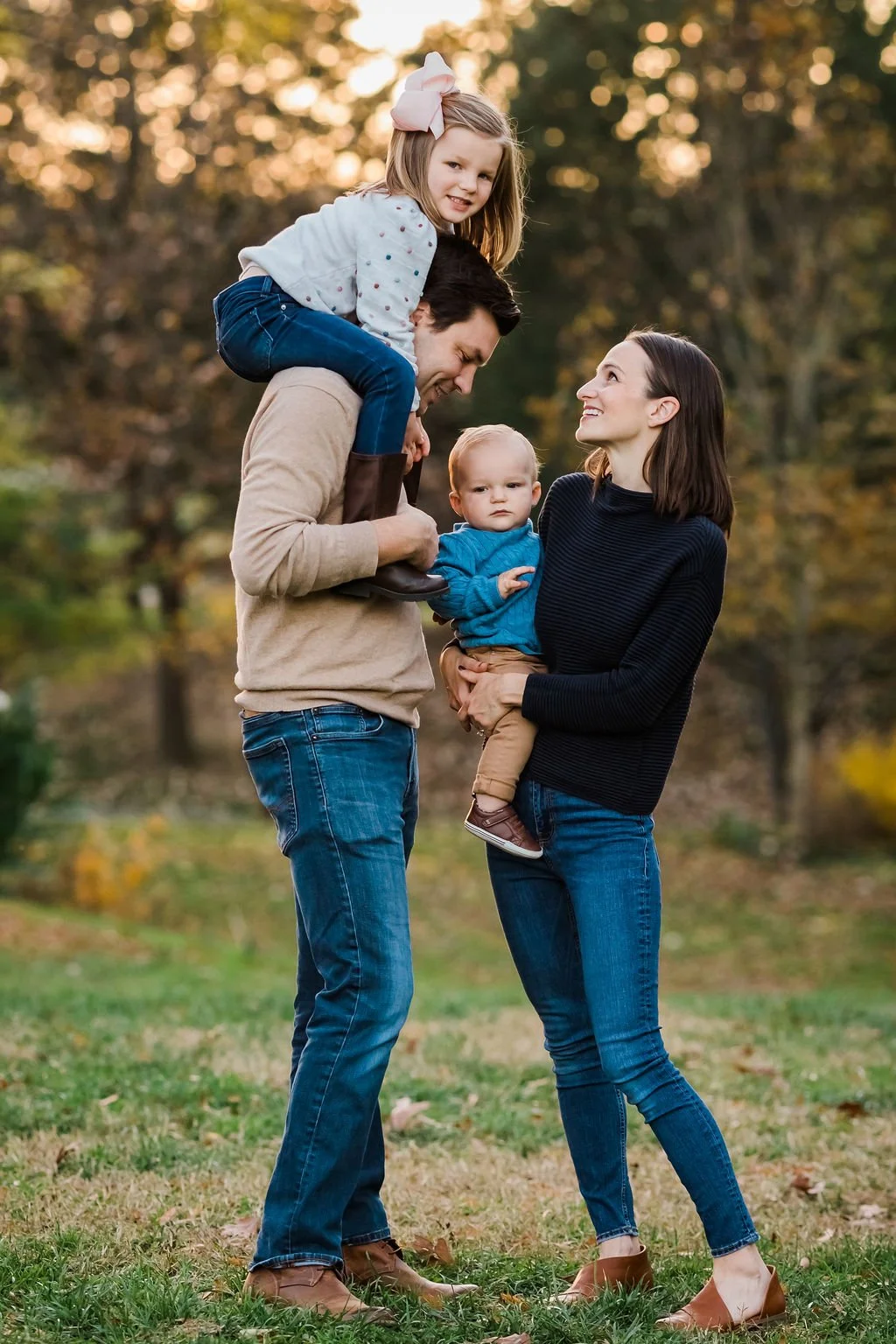 A family of four outdoors during autumn, with a man carrying a young girl on his shoulders and a woman holding a toddler, all smiling and looking at each other.