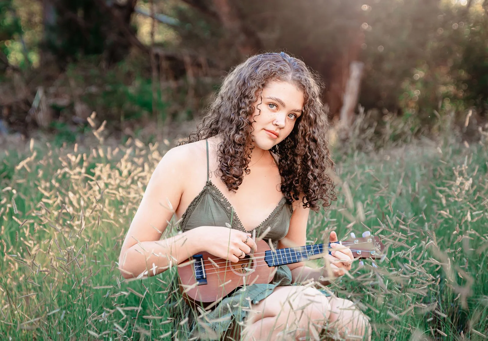 A young woman with curly hair sitting in a field of tall grass, playing a small ukulele. She wears a green tank top and shorts, looking at the camera with a gentle expression. The background shows trees and a soft, natural light.