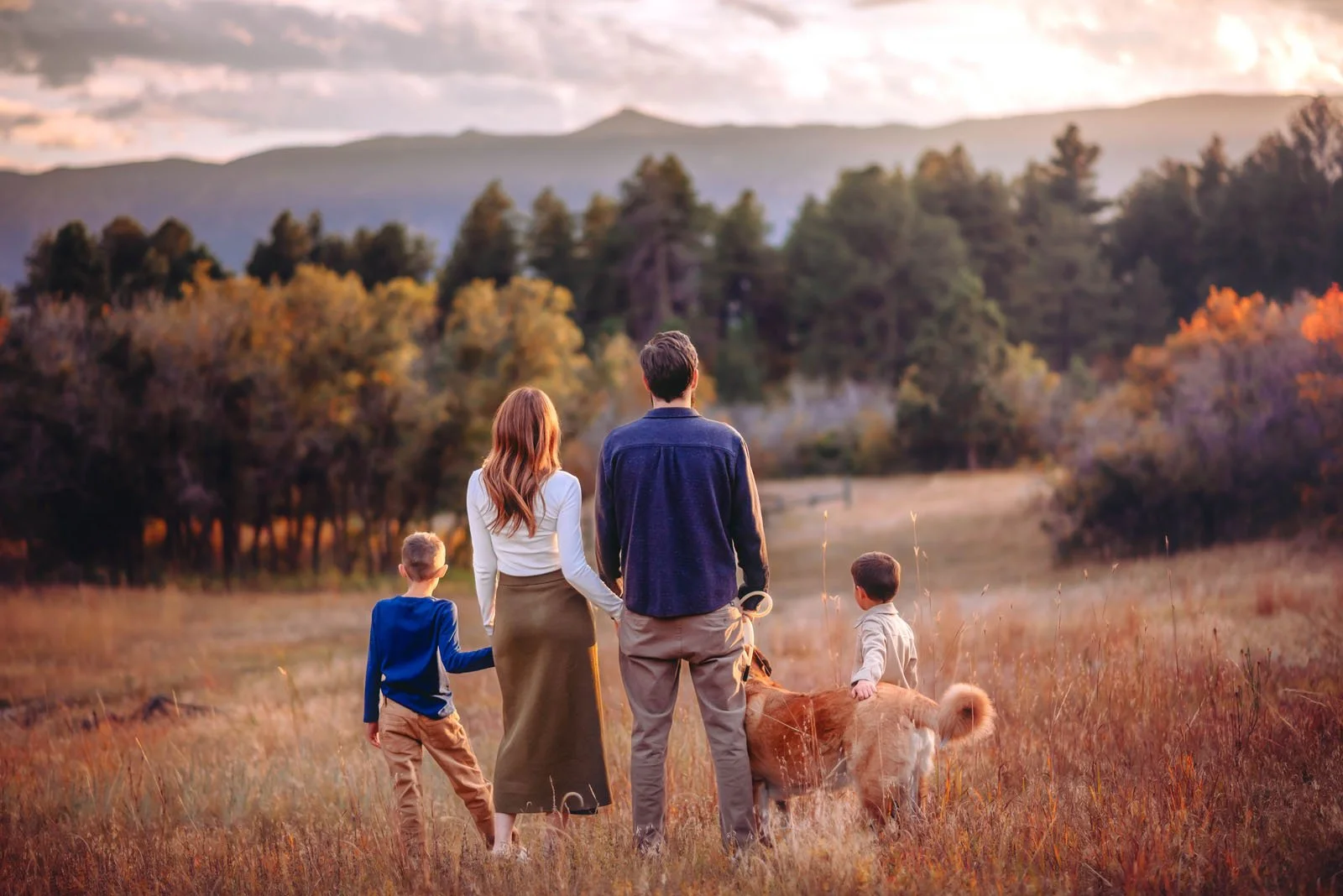 A family of four, including two children, a woman, and a man, walks with a dog through a meadow with tall grass, trees, and mountains in the background during sunset.