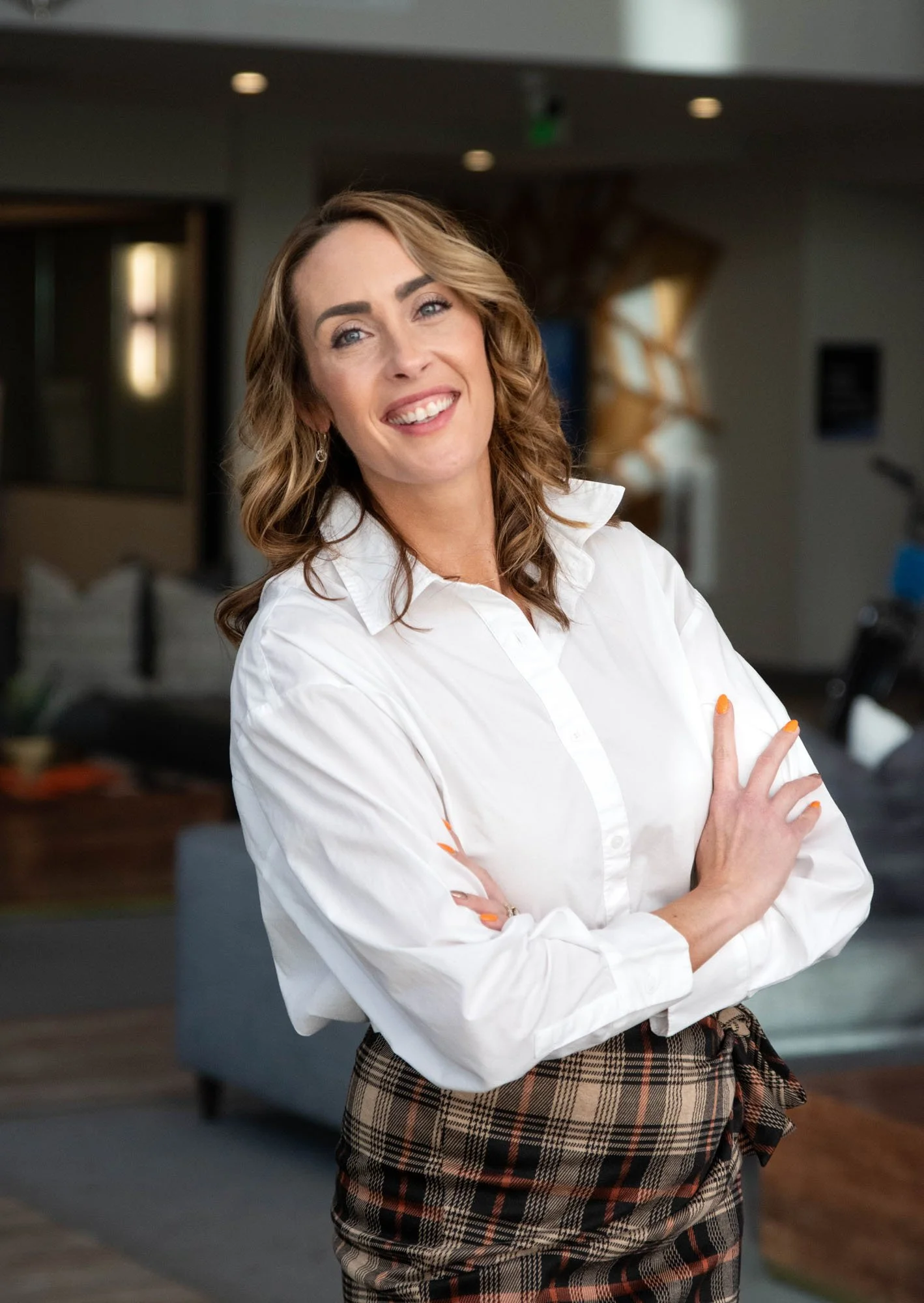 A woman with wavy brown hair, blue eyes, and light makeup, smiling and crossing her arms, wearing a white button-up shirt and a brown plaid skirt with a matching plaid sash, standing indoors with a blurred background.