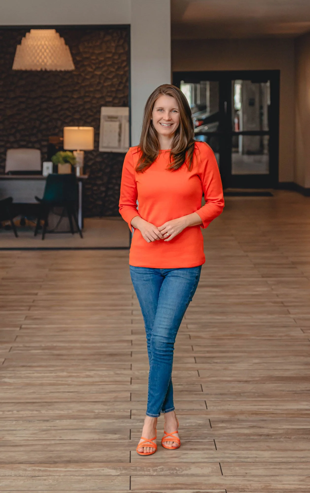 A smiling woman with long brown hair wearing an orange long-sleeve top, blue jeans, and orange strappy heels standing on a wooden floor in an indoor space with a modern, cozy decor background.