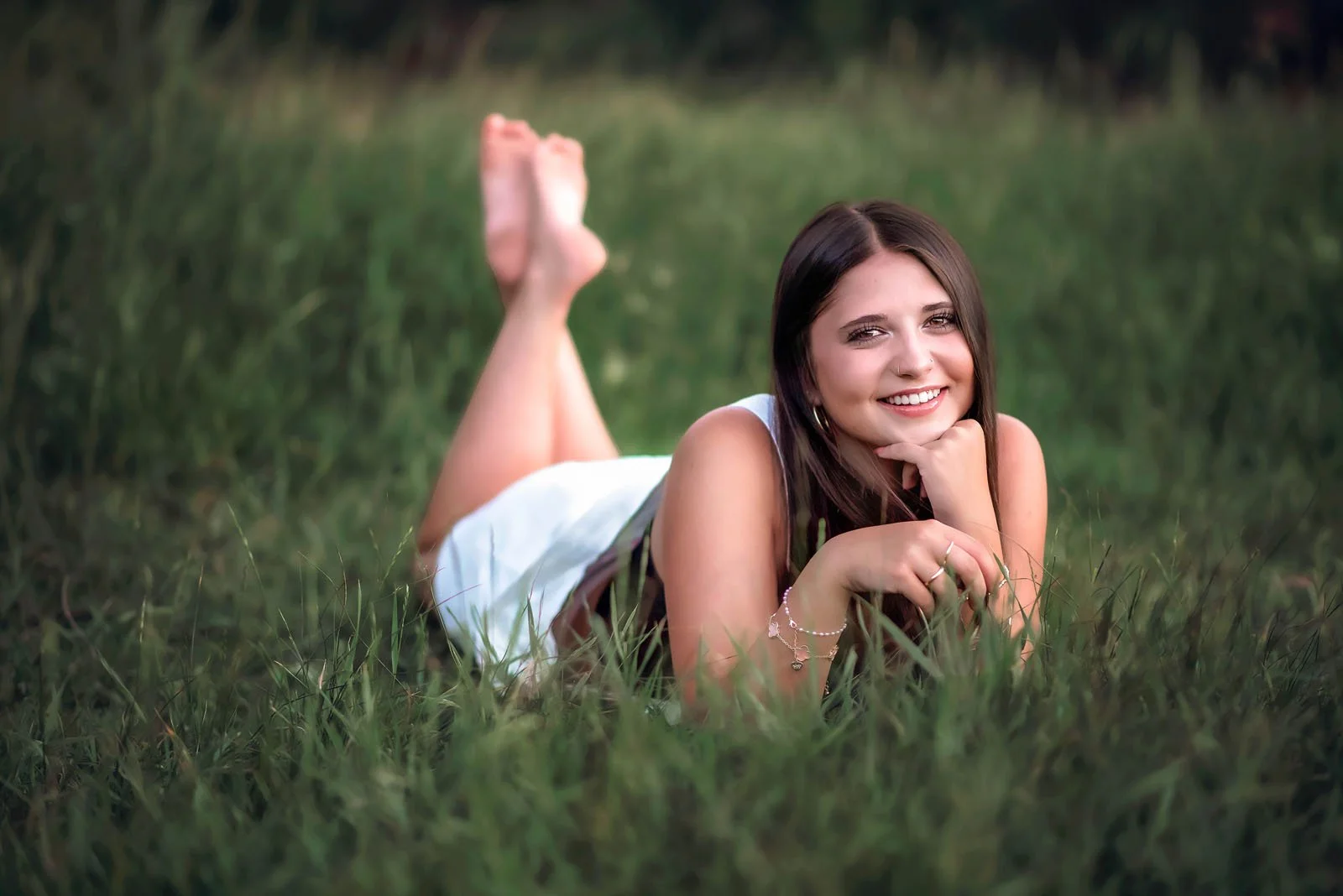 Young woman with long dark hair lying on her stomach in the grass, smiling at the camera with her head resting on her hand, wearing a white top and jewelry.