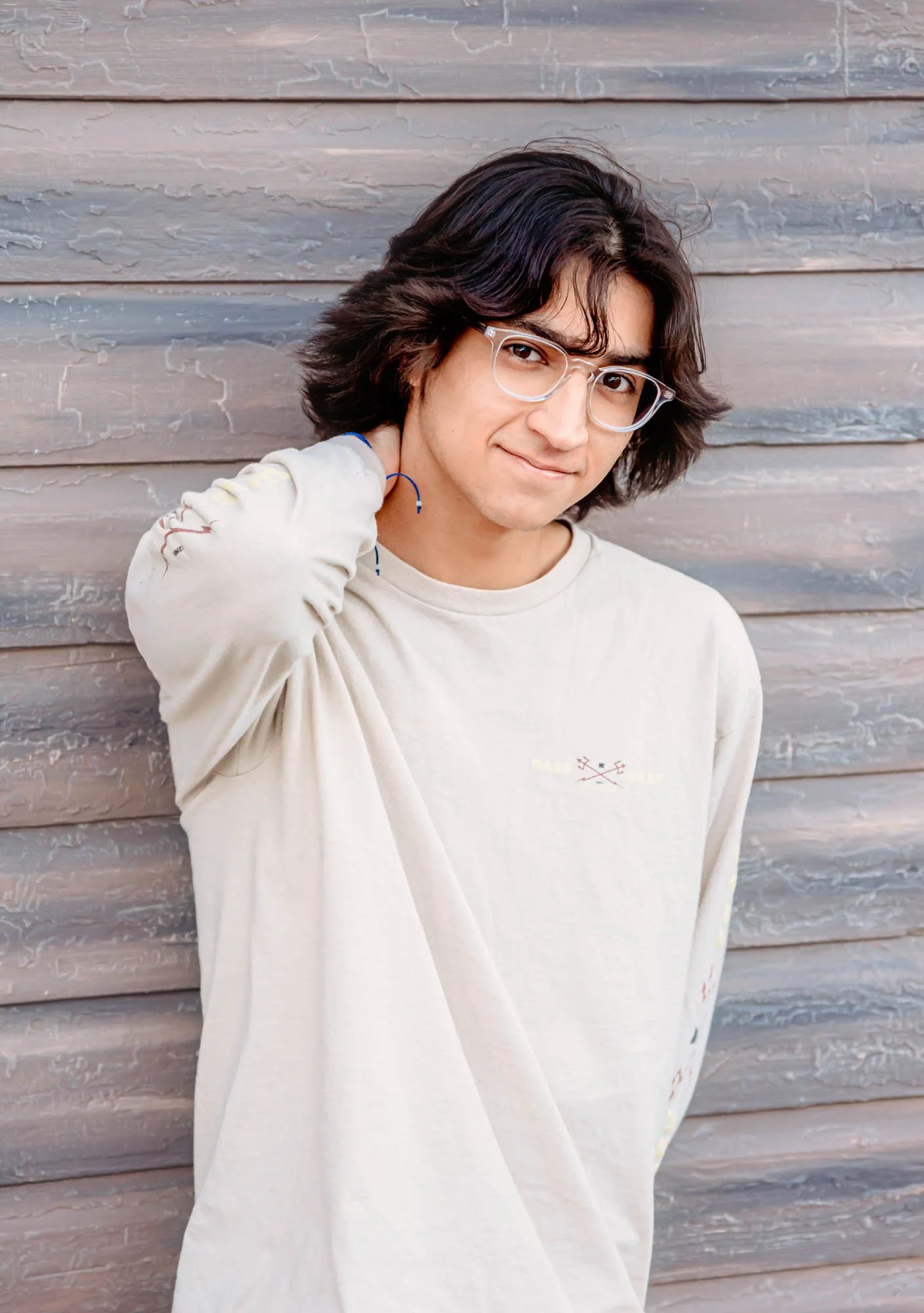 A young man with medium-length dark hair, clear glasses, and a light-colored sweatshirt is standing against a weathered wooden wall, smiling gently and looking into the camera.